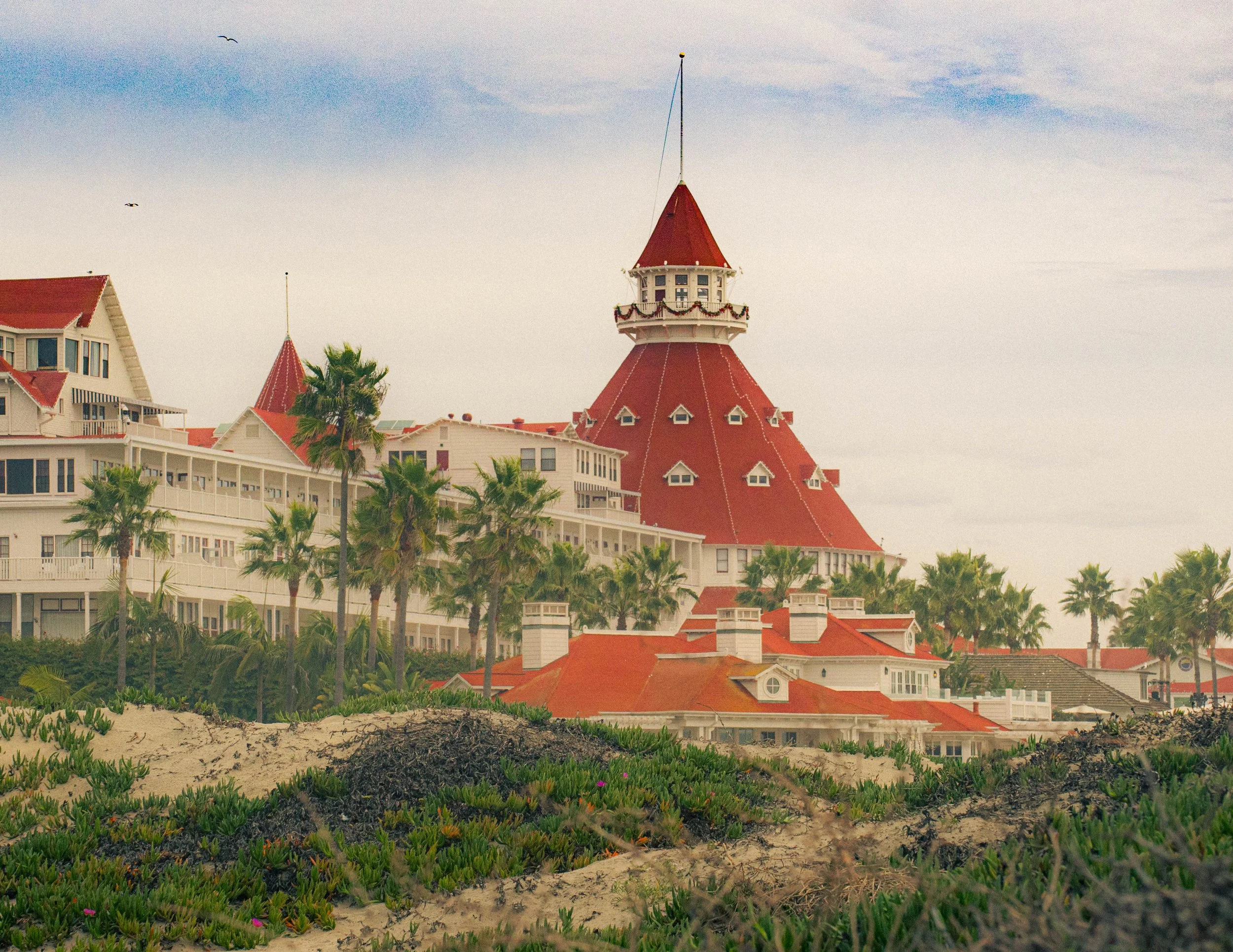 A seaside resort with white buildings and red rooftops, green bushes, palm trees, and a sandy area in the foreground, under a partly cloudy sky with seagulls flying.