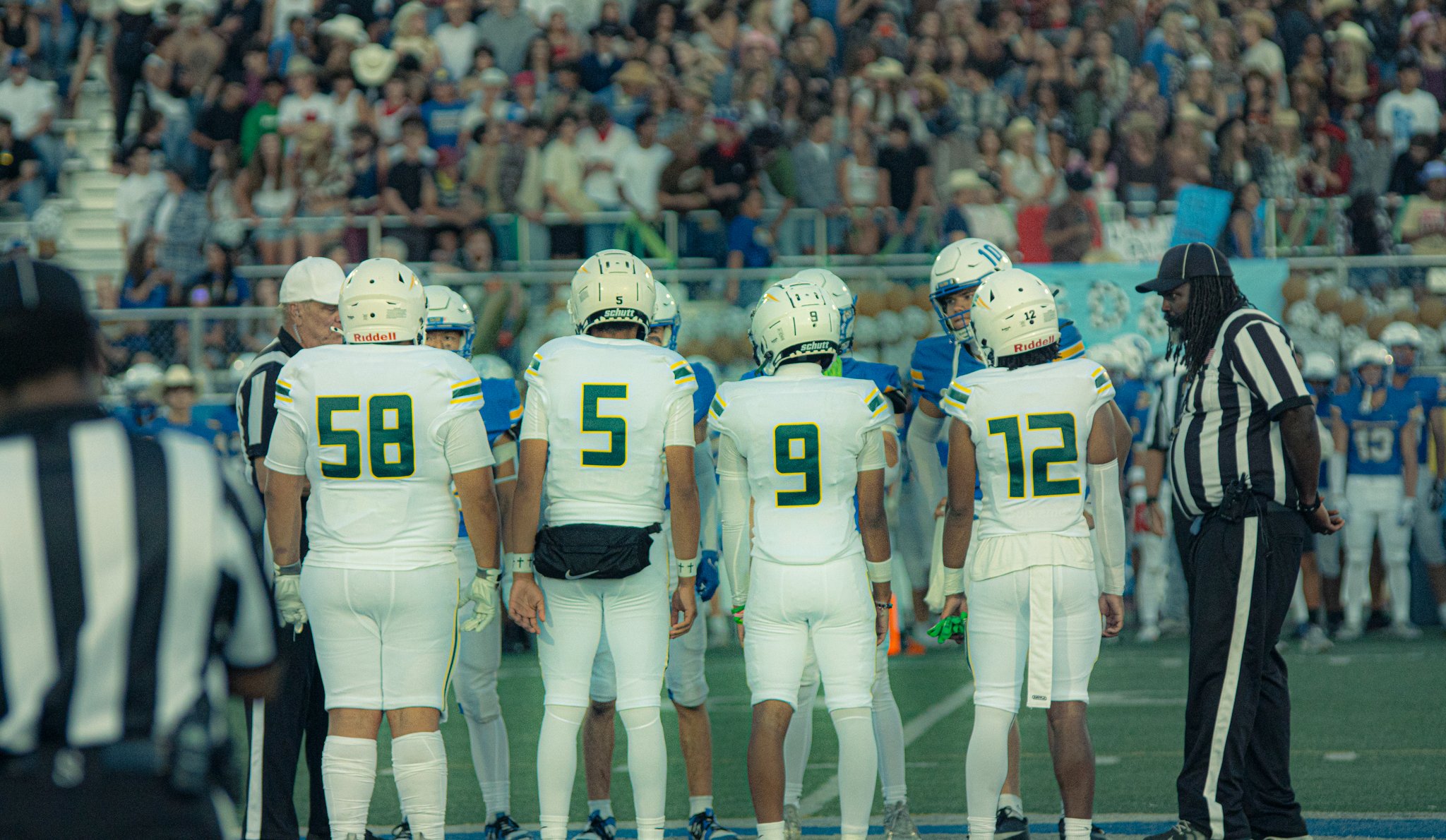 A football team wearing white jerseys and helmets gather on the field during a game, with referees nearby and a crowd of spectators in the stands.