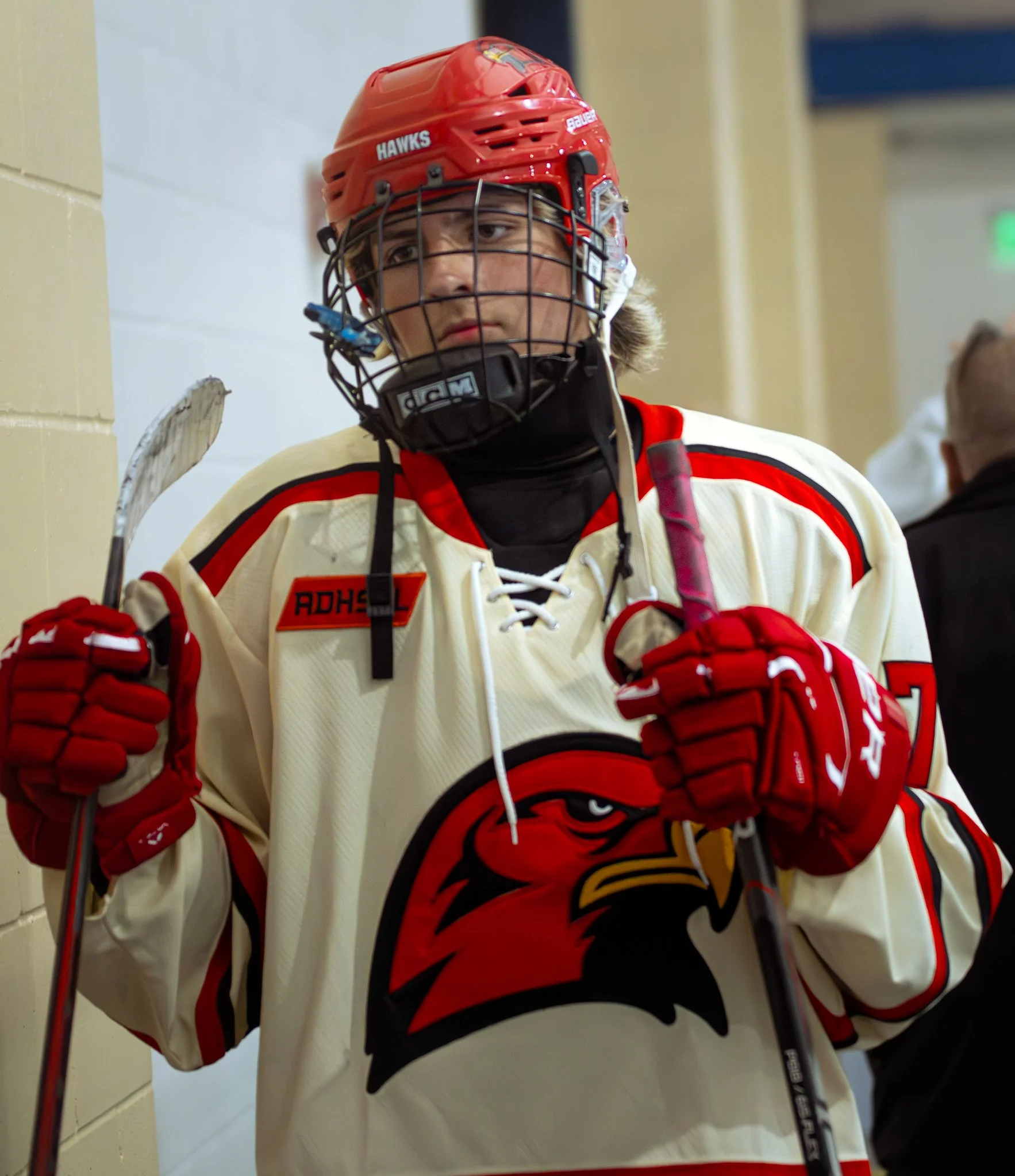 Close-up of a hockey player wearing a red helmet with a black cage mask, a white and red jersey with a tiger logo, and red gloves, holding a hockey stick, in an indoor setting.