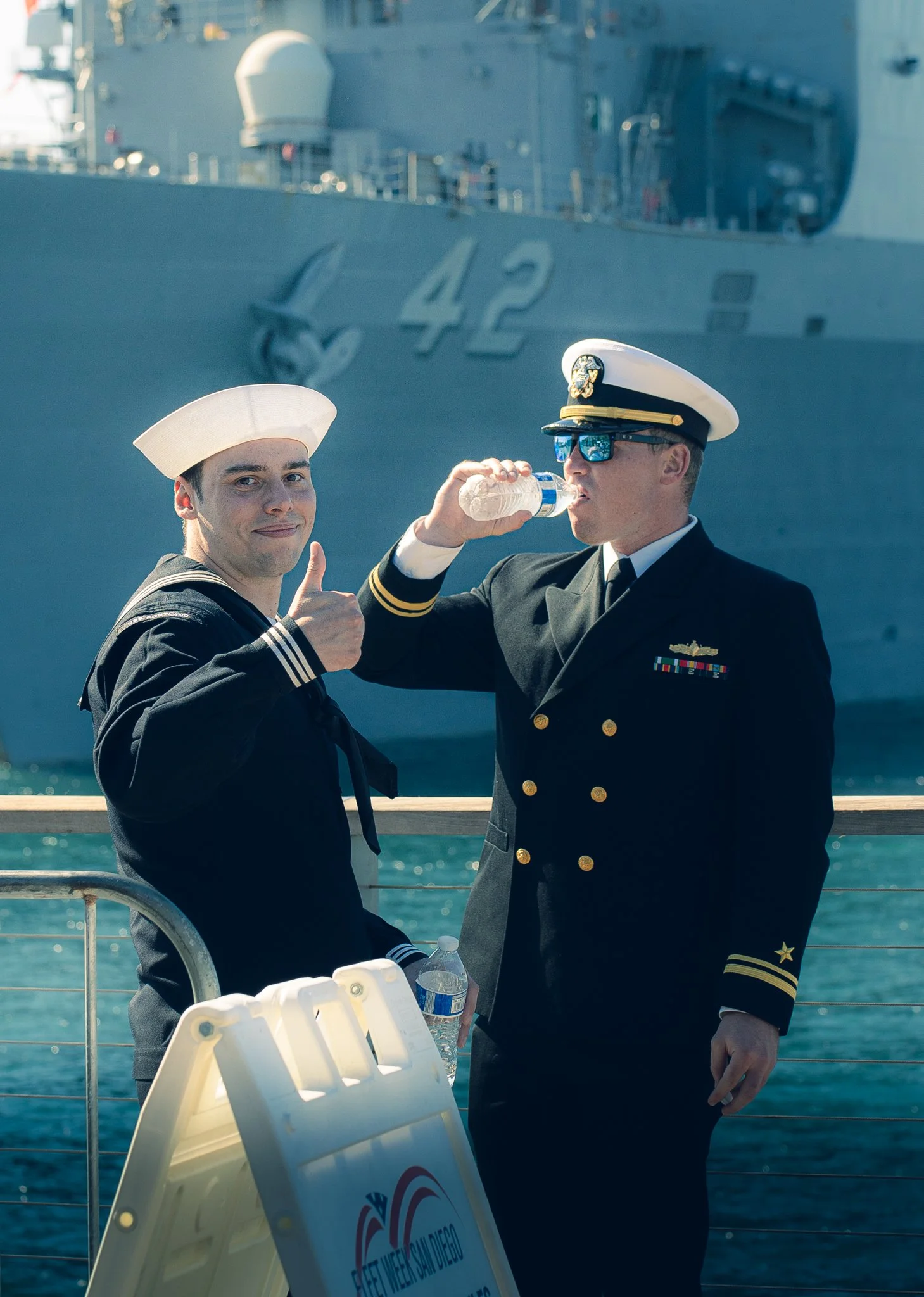 Two sailors in uniform on a ship deck, one wearing a sailor hat and smiling with a thumbs-up, the other in a navy uniform drinking water, with a large naval ship in the background.