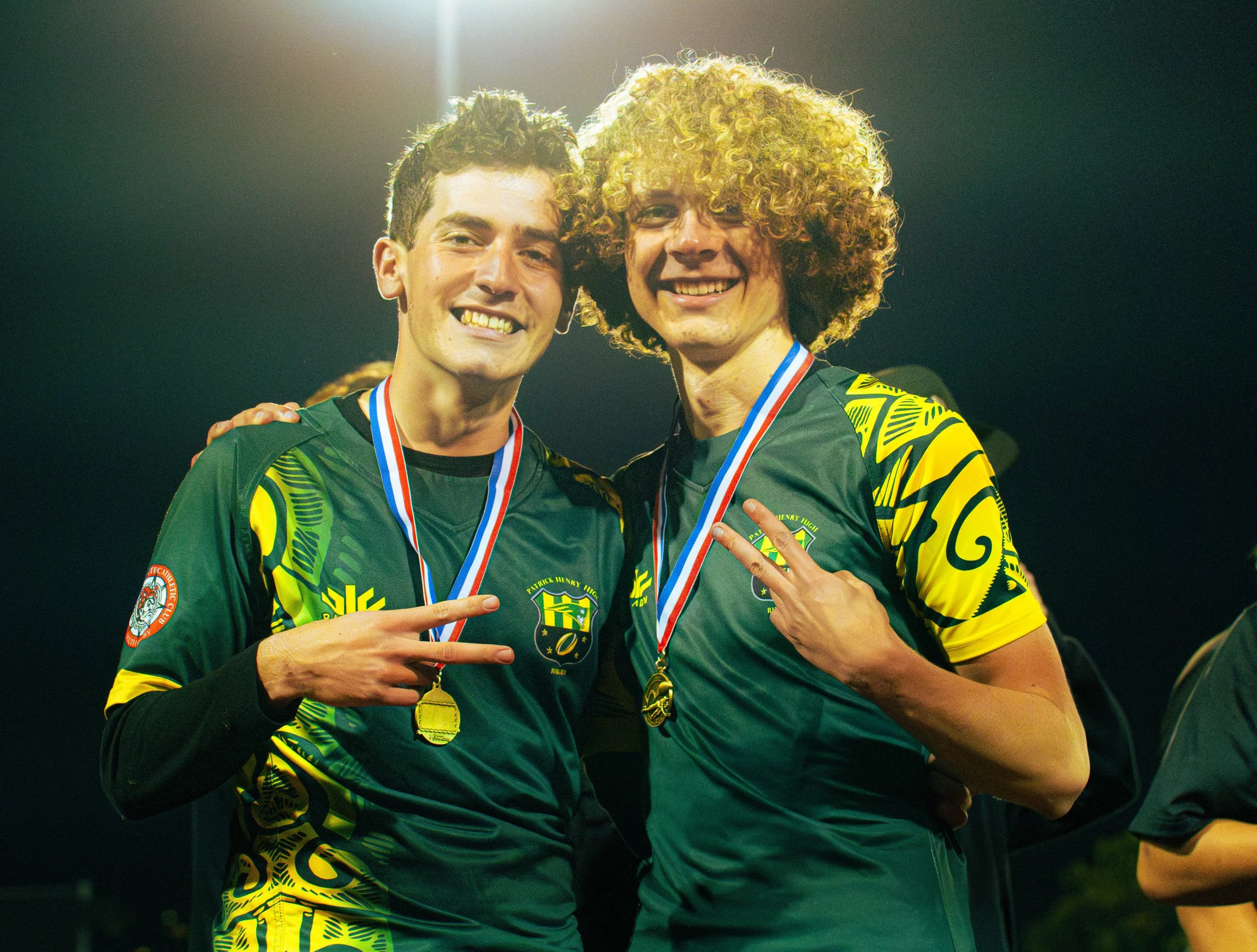 Two young men celebrating with medals around their necks, wearing green and yellow sports jerseys, making victory signs with their hands, smiling, in a brightly lit environment.