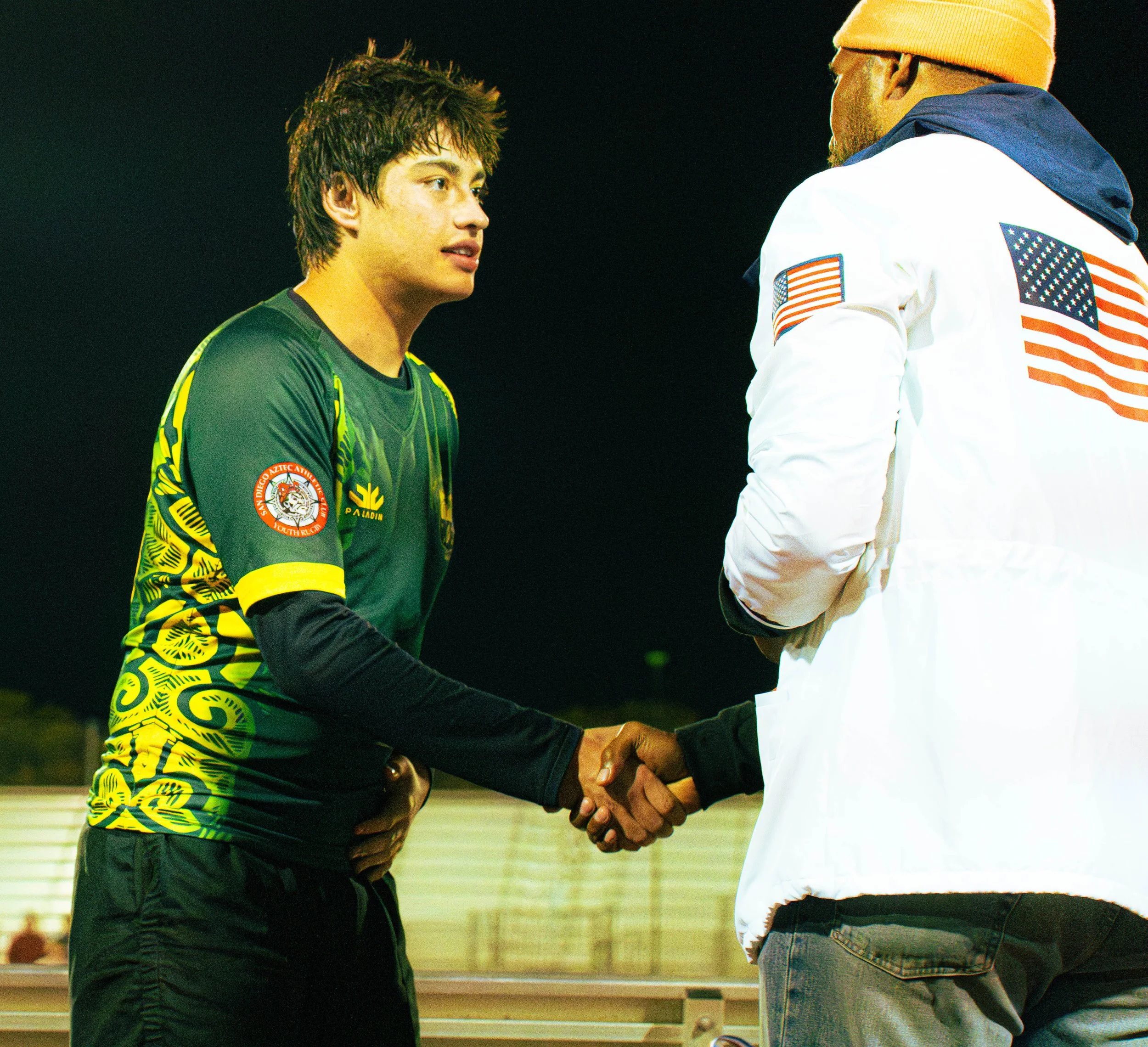 Two men shaking hands indoors, one with dreadlocks and a green shirt, the other wearing a beige beanie and a white jacket with American flags on the sleeves.