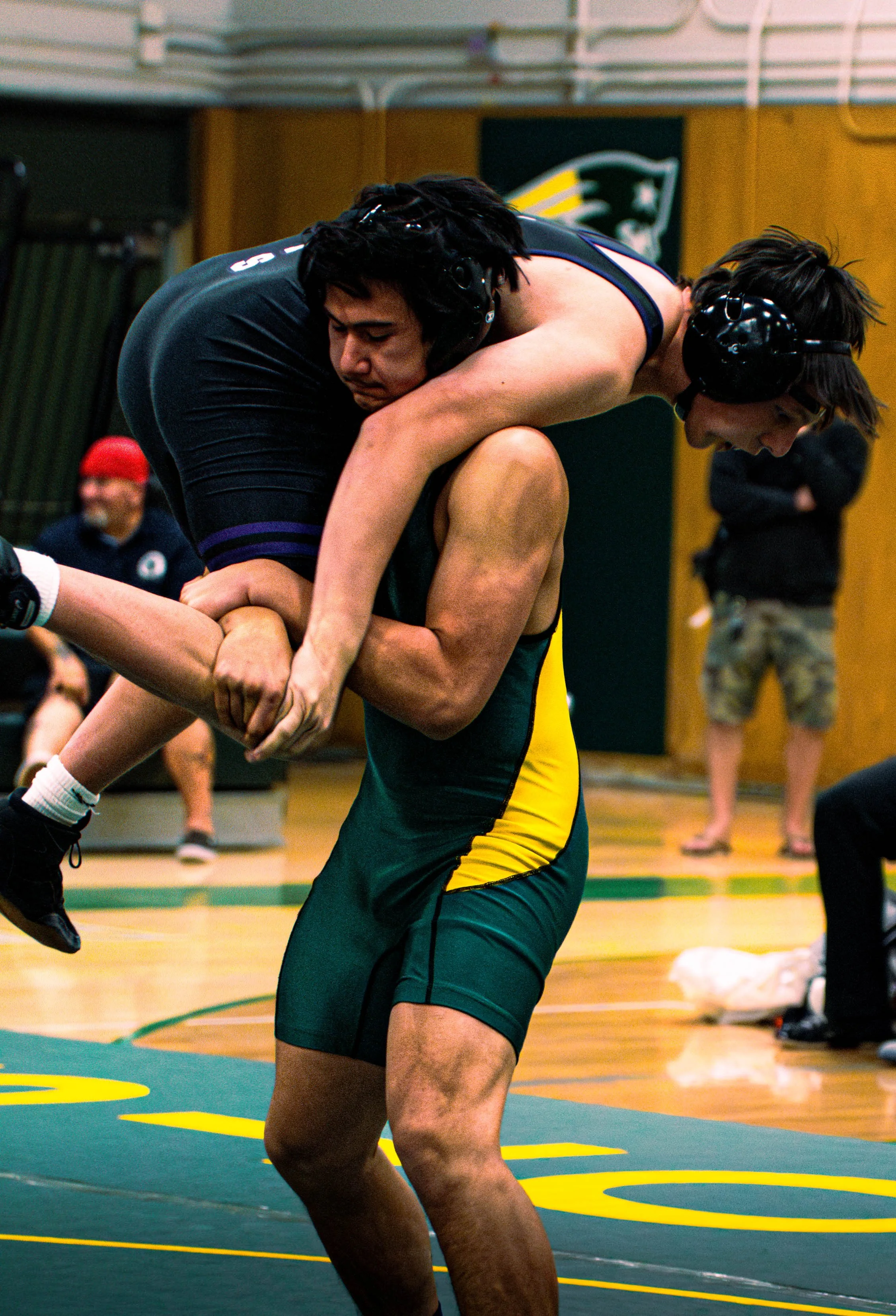 Two male wrestlers engaging in a match on a wrestling mat with yellow rings, wearing protective headgear and athletic uniforms, in a gymnasium with spectators and a flag in the background.