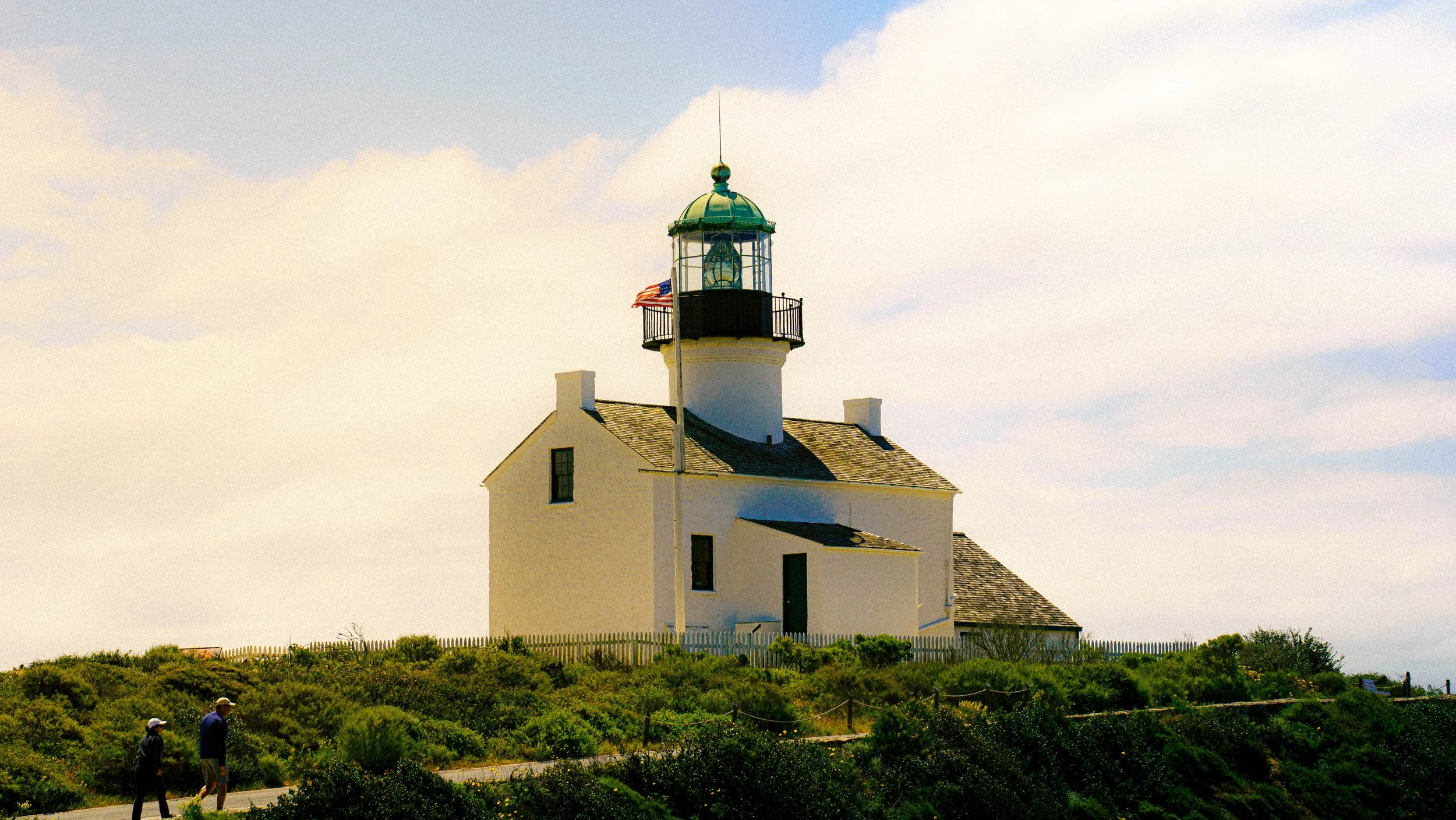 A white lighthouse with a green metal roof and a small balcony, flying an American flag, stands against a cloudy sky. There are bushes and a walking path with two people in the foreground.