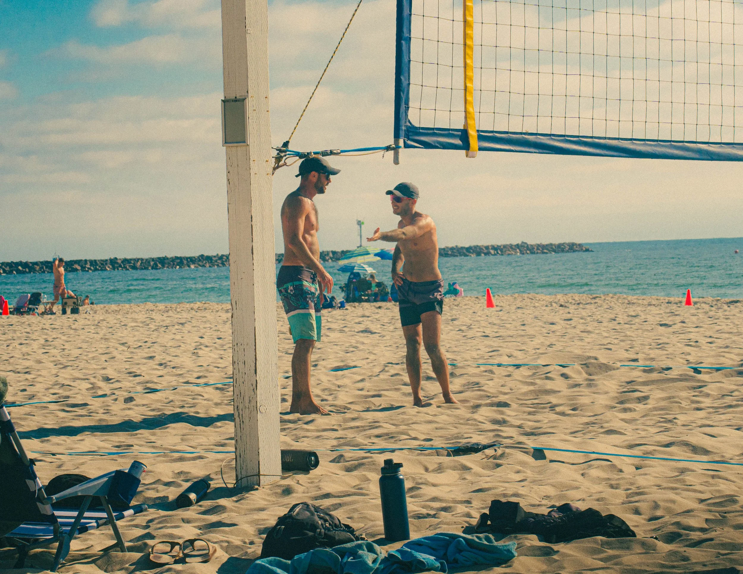 Two men are playing beach volleyball near the ocean, with one gesturing and the other listening. Beach chairs, umbrellas, and personal items are scattered on the sand, with a breakwater and a few people in the background. It is a sunny day with a par