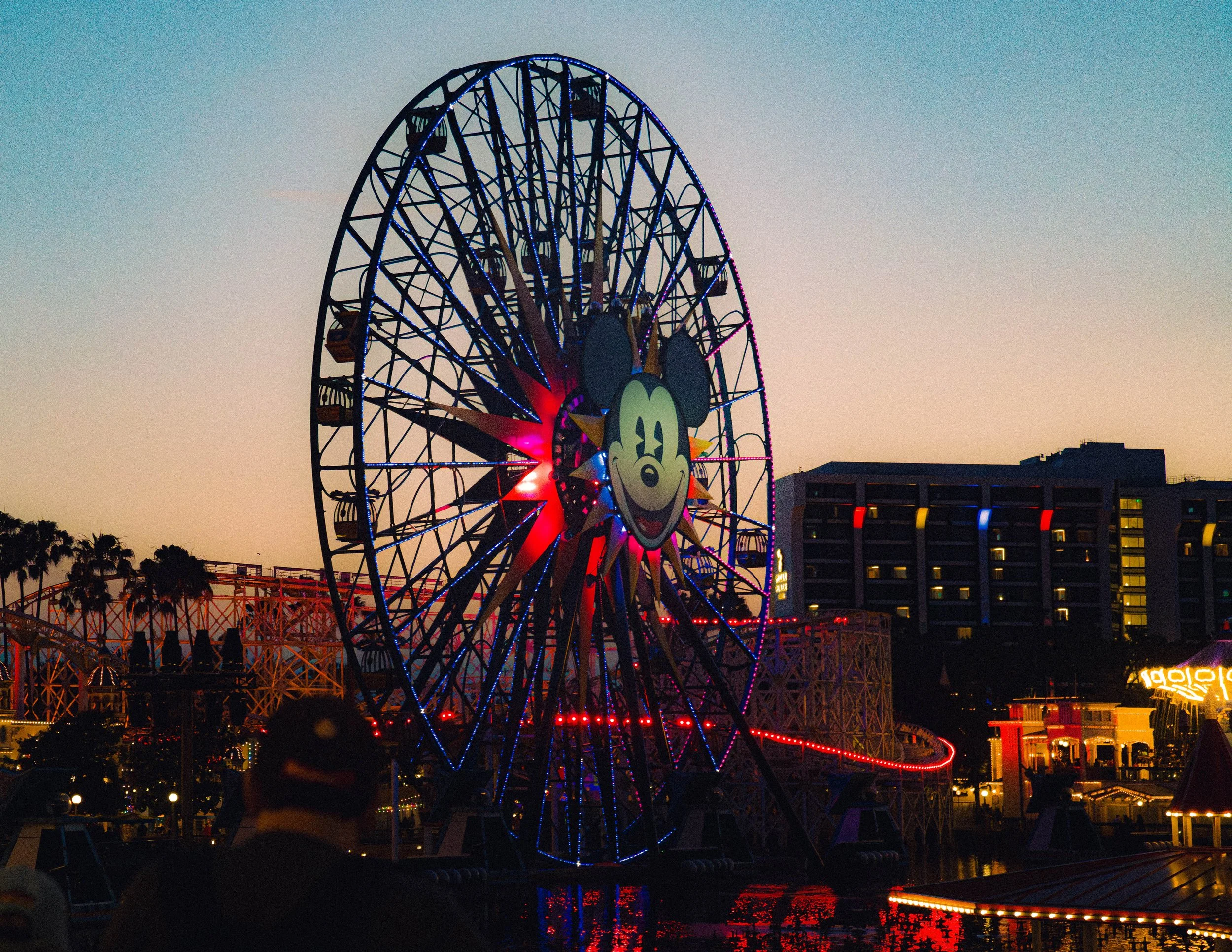 A Ferris wheel decorated with colorful lights at dusk, featuring a Mickey Mouse face at the center, with a roller coaster track and building with illuminated windows in the background.