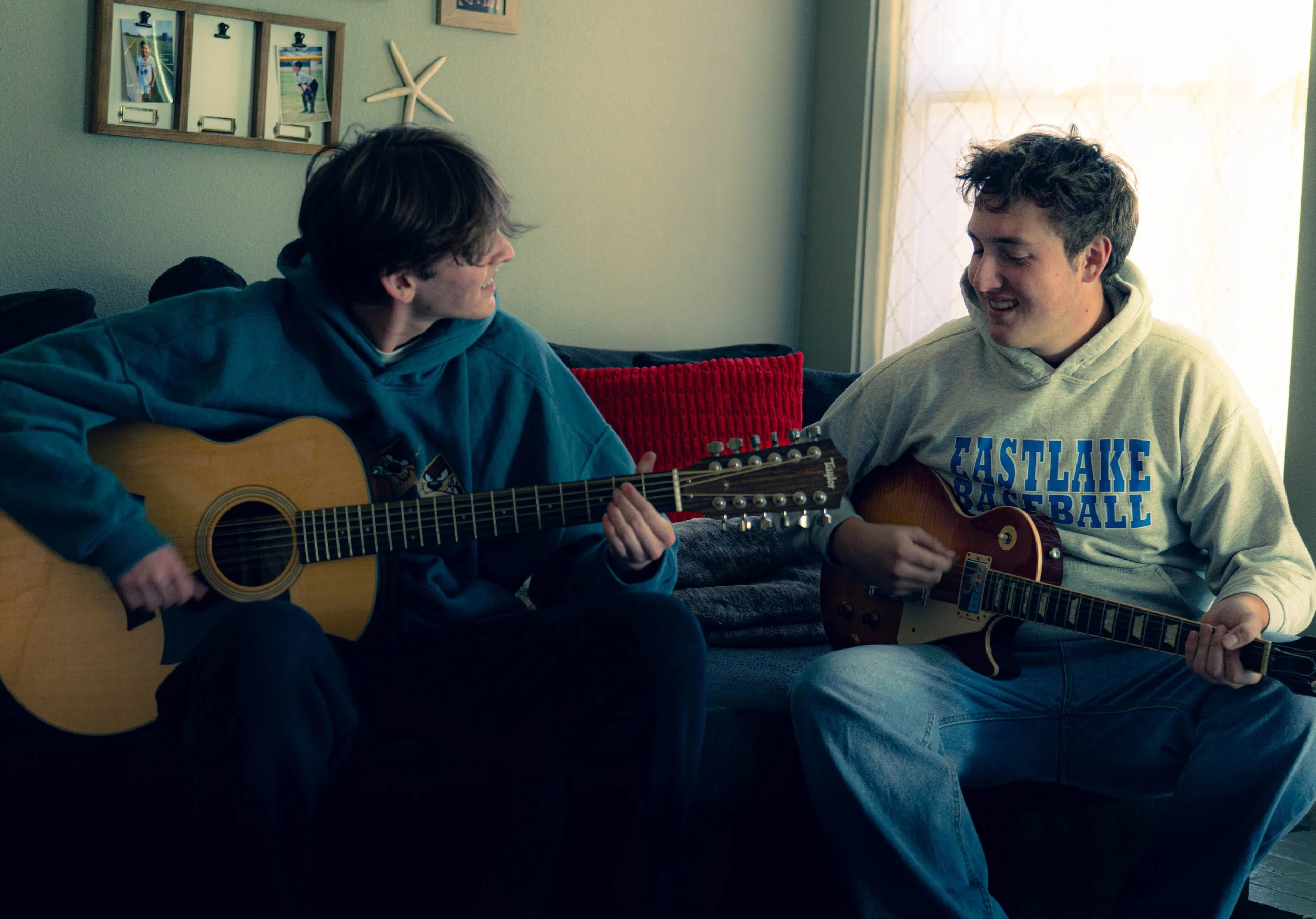 Two young men sitting on a sofa, playing acoustic guitars and sharing a moment of music and friendship in a cozy, well-lit living room.