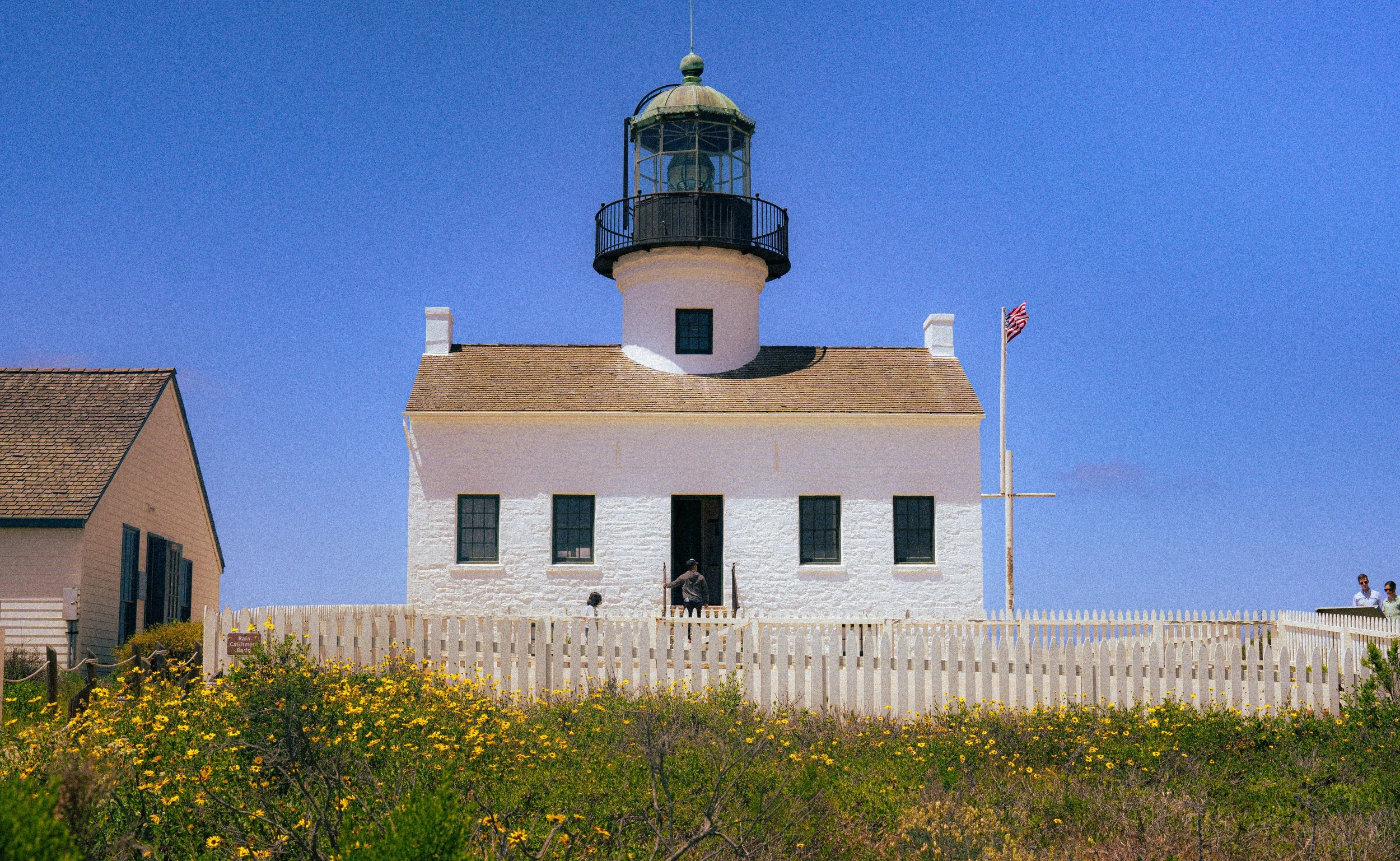 A lighthouse with a white tower and a black lantern room on top, surrounded by a white picket fence, with a blue sky in the background. There are a few people near the entrance and an American flag flying on a flagpole to the right.