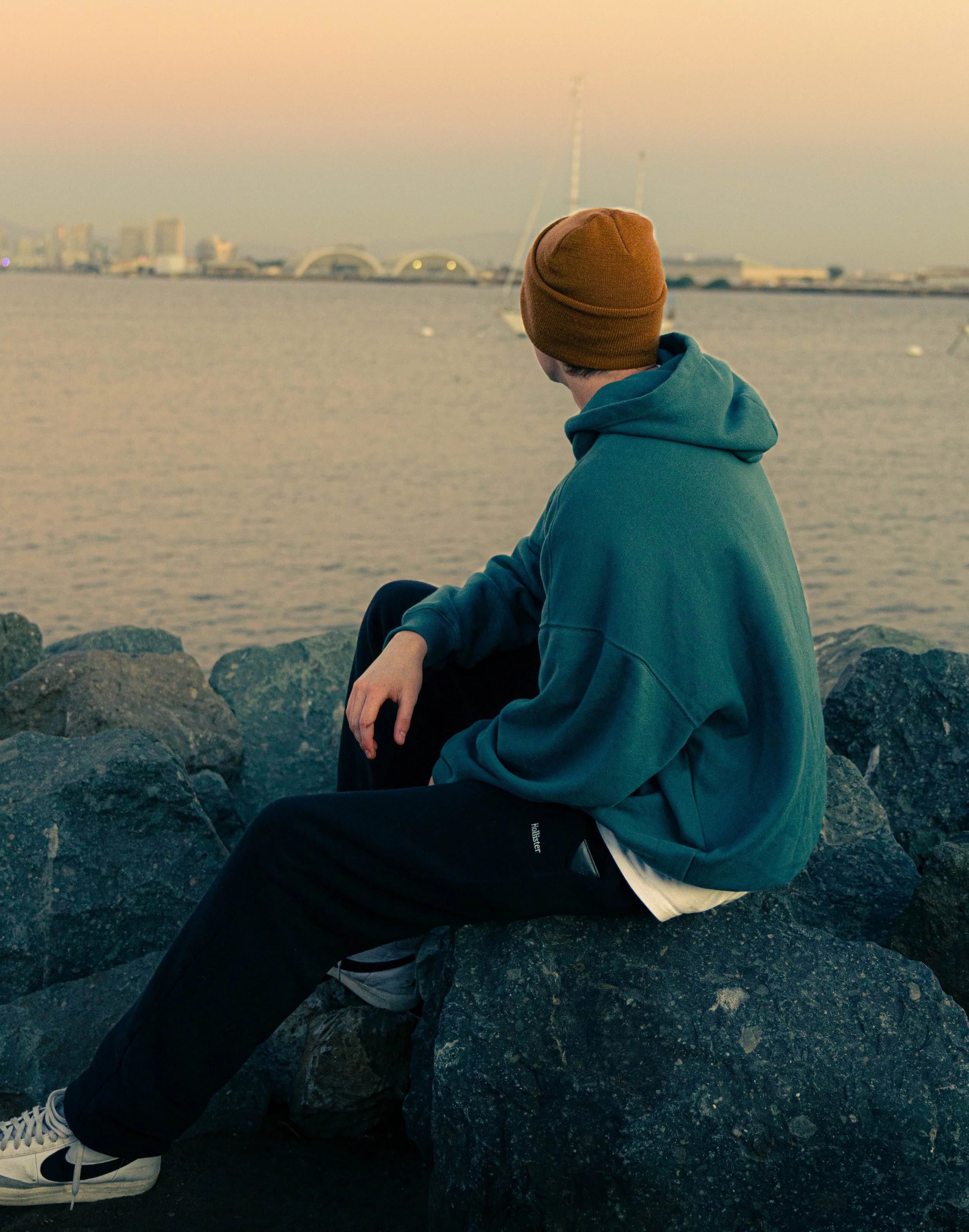 Person sitting on rocks by the water, wearing a brown beanie, teal hoodie, black pants, and white sneakers, looking towards the city skyline with sailboats in the water.