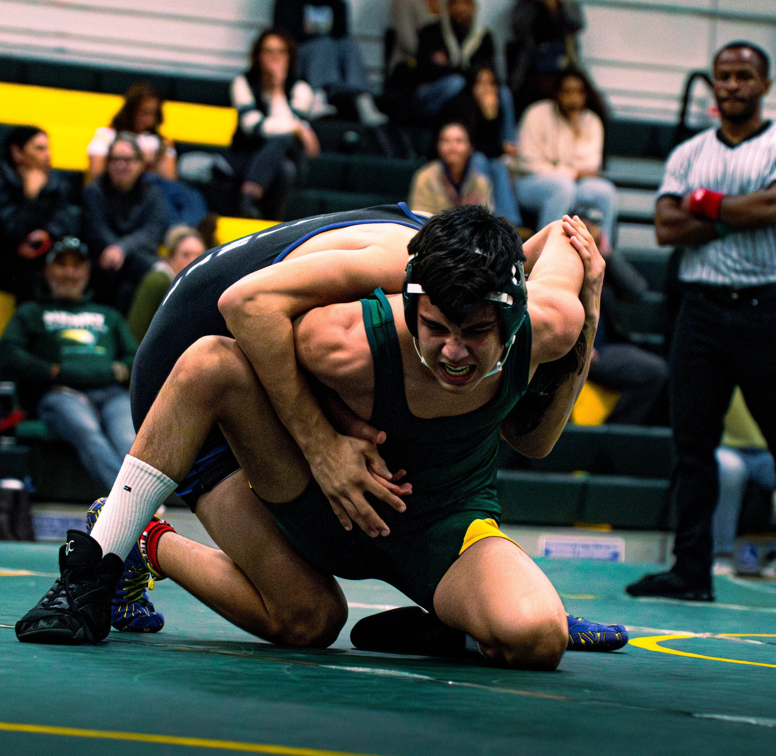 Two wrestlers compete on a wrestling mat with one wrestler pinning the other down. Spectators are seated in the background watching the match.