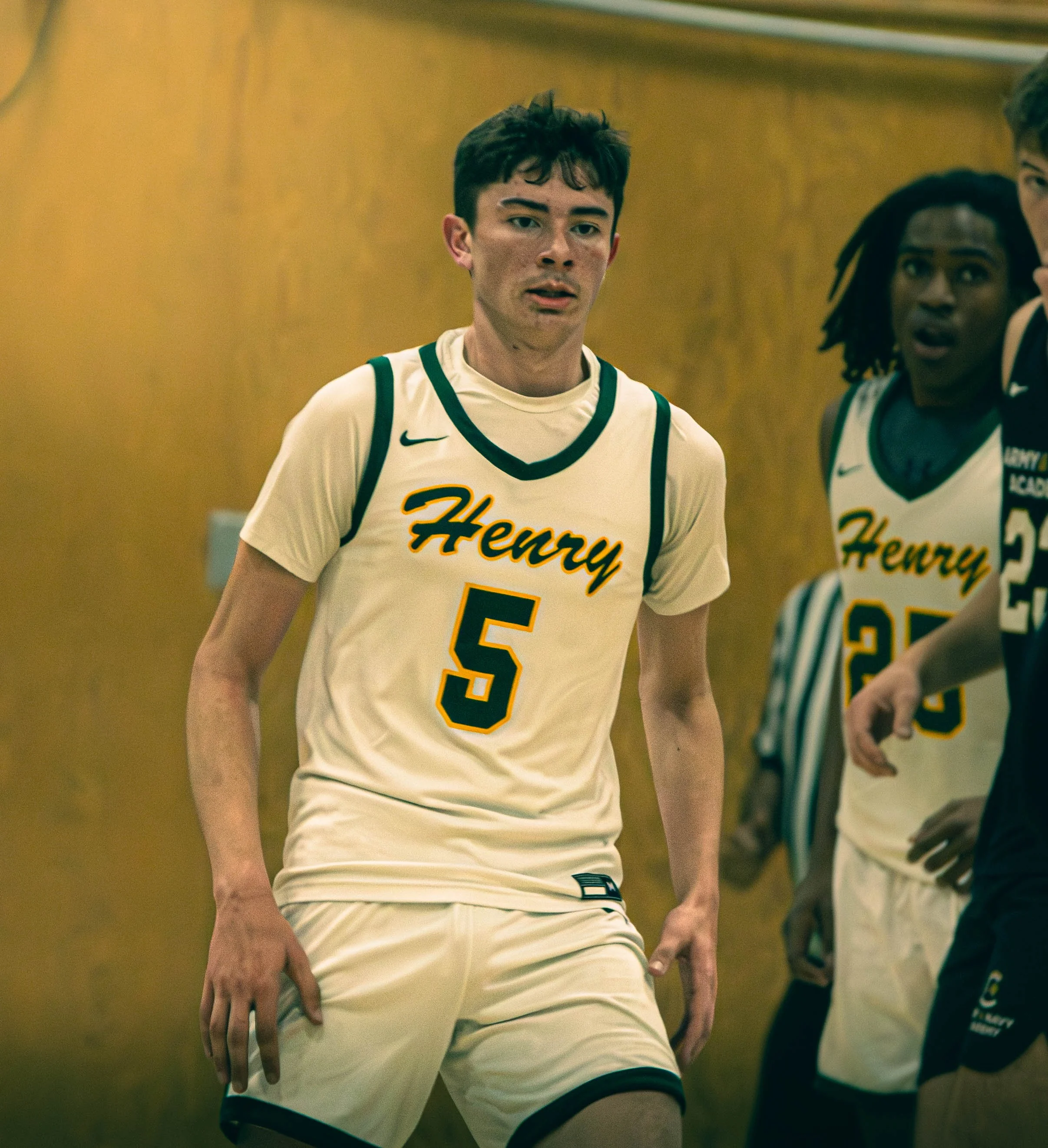 A young male basketball player in a cream-colored uniform with green accents and the number 5, standing in a gymnasium with team members behind him. The gym wall is wood-paneled.
