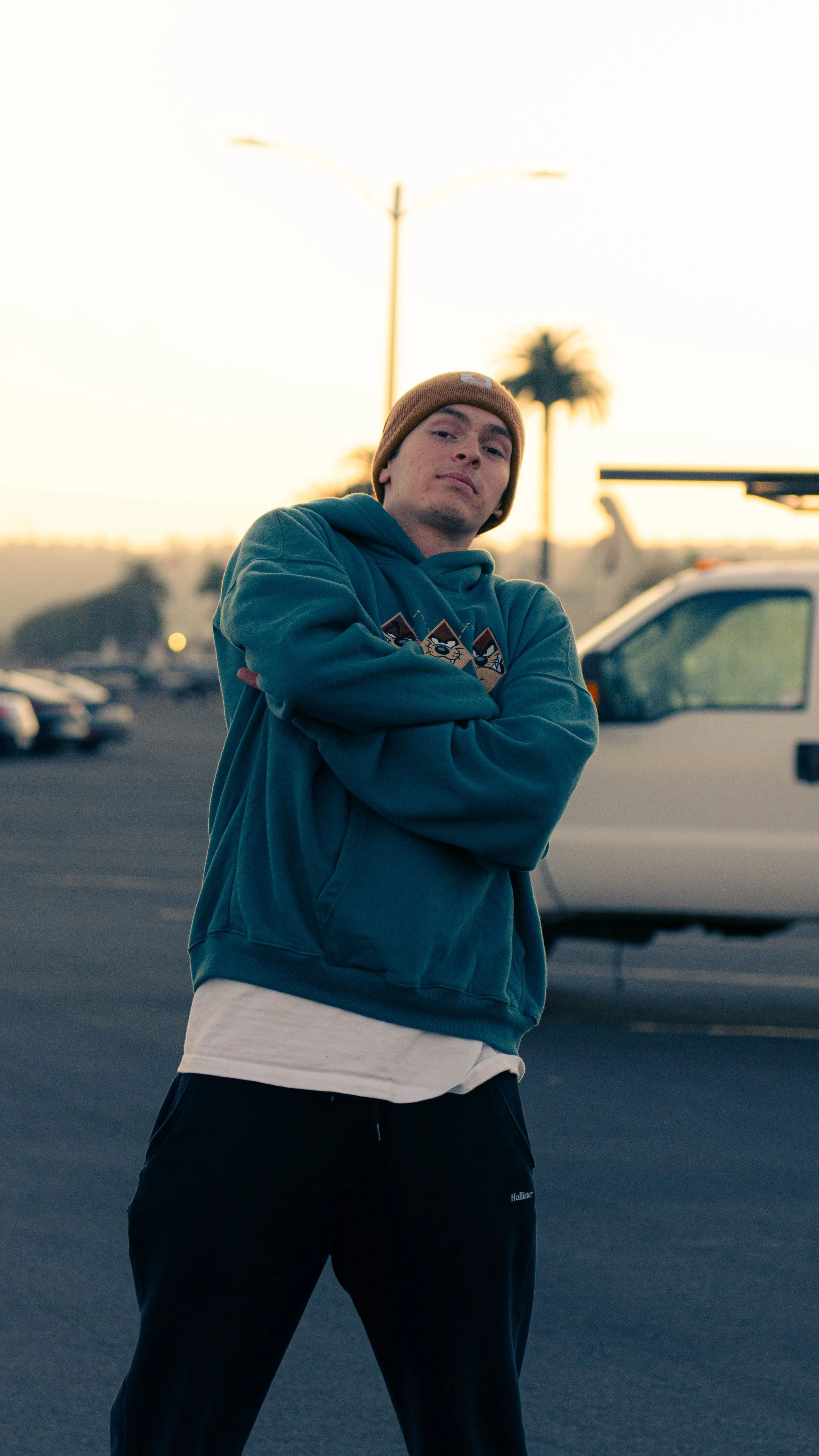 A young man stands outdoors in a parking lot with his arms crossed, wearing a brown beanie, teal hoodie with a graphic design, and dark pants, with parked cars and palm trees in the background at sunset.