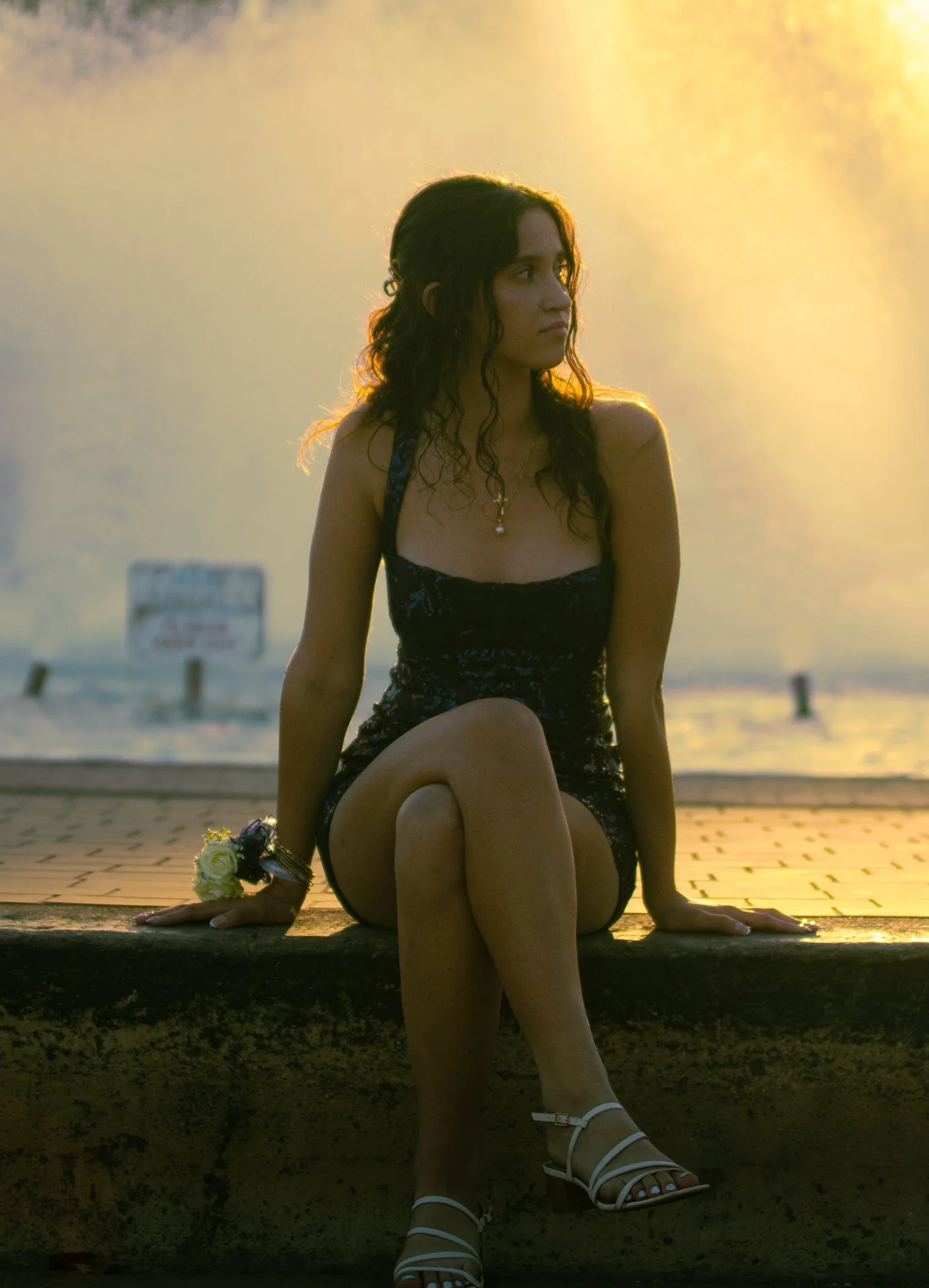 A young woman sitting on a ledge near the water during sunset, wearing a dark dress, white heels, and a flower bracelet, looking to the right.