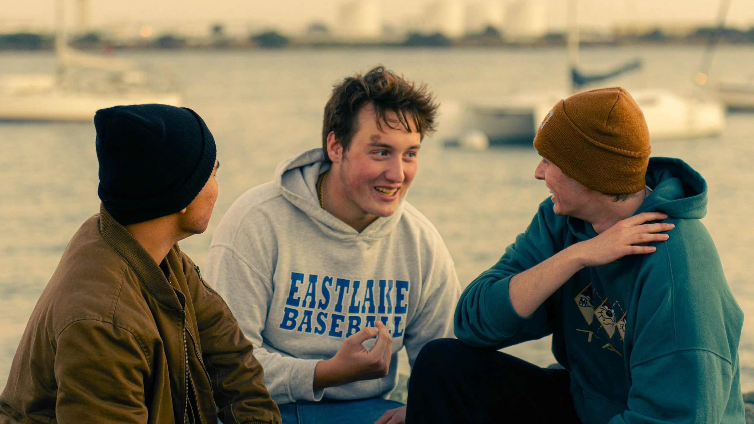 Three young men sitting outdoors by the water, engaged in conversation, with boats and a harbor in the background.