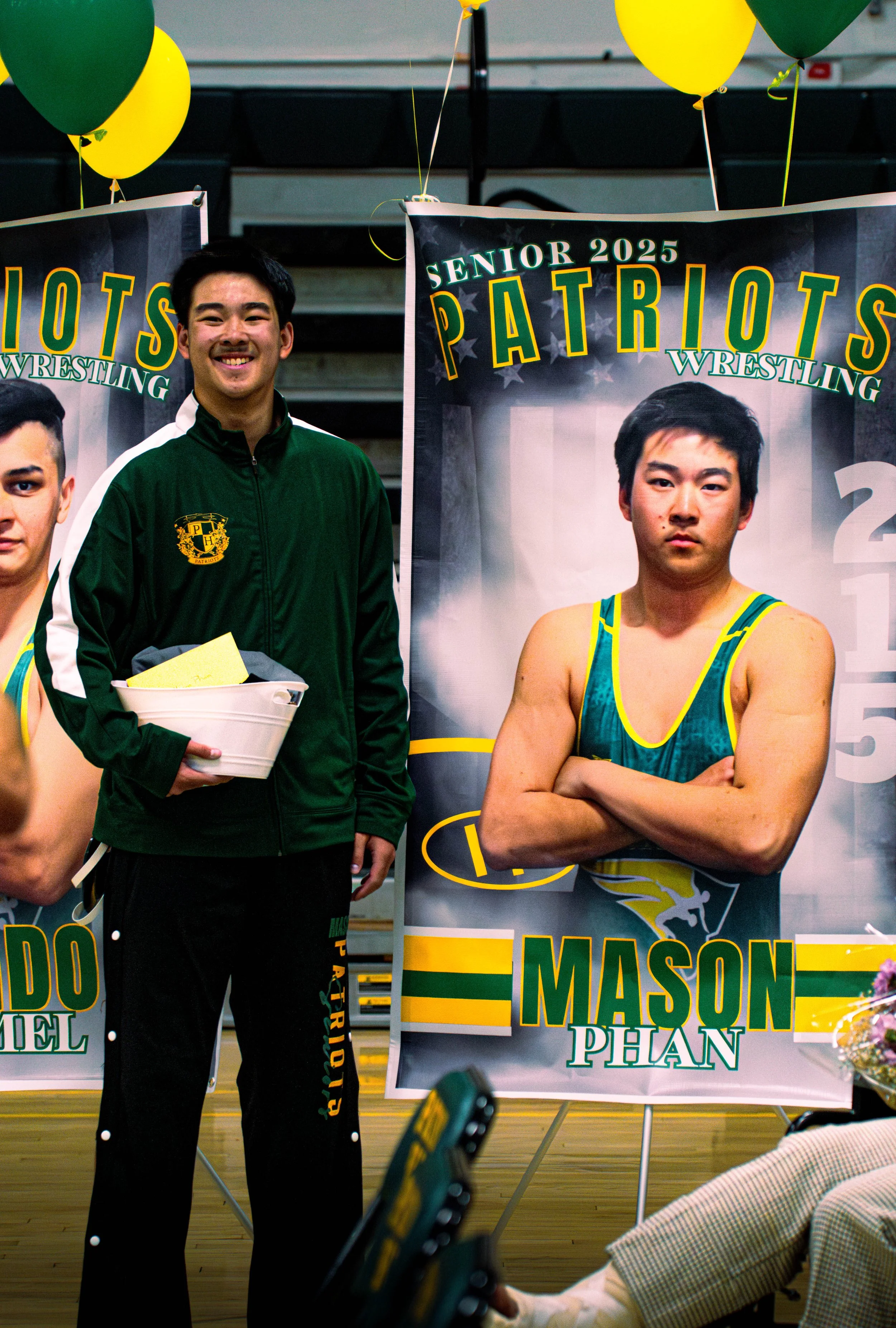 Young man in a green jacket and black pants standing beside a podium with a poster of a wrestler named Mason Phan, with balloons and other people in the background at a wrestling event.
