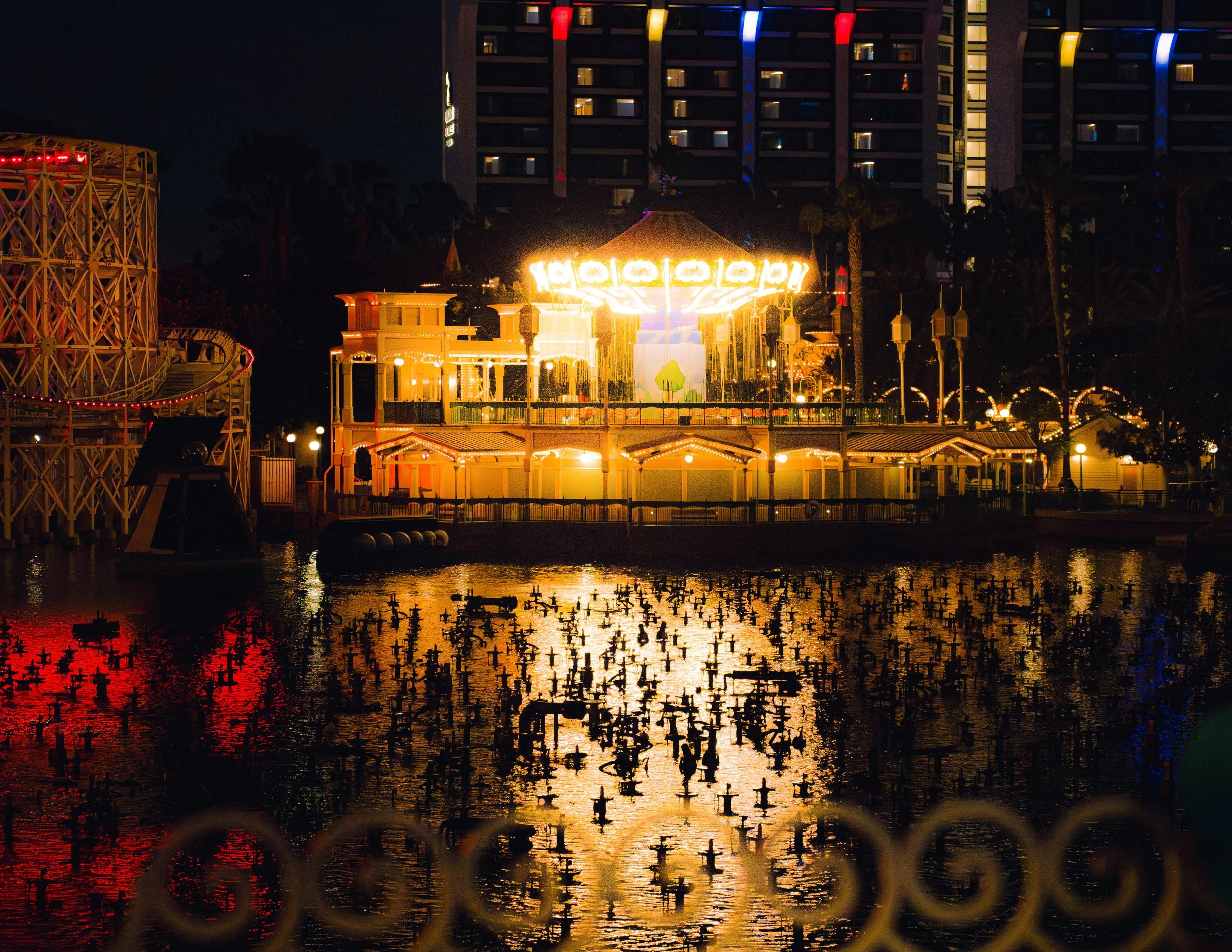Night scene of a brightly lit amusement park ride with a large circular structure and colorful lights, reflected on water below, with a multi-story hotel or apartment building in the background.
