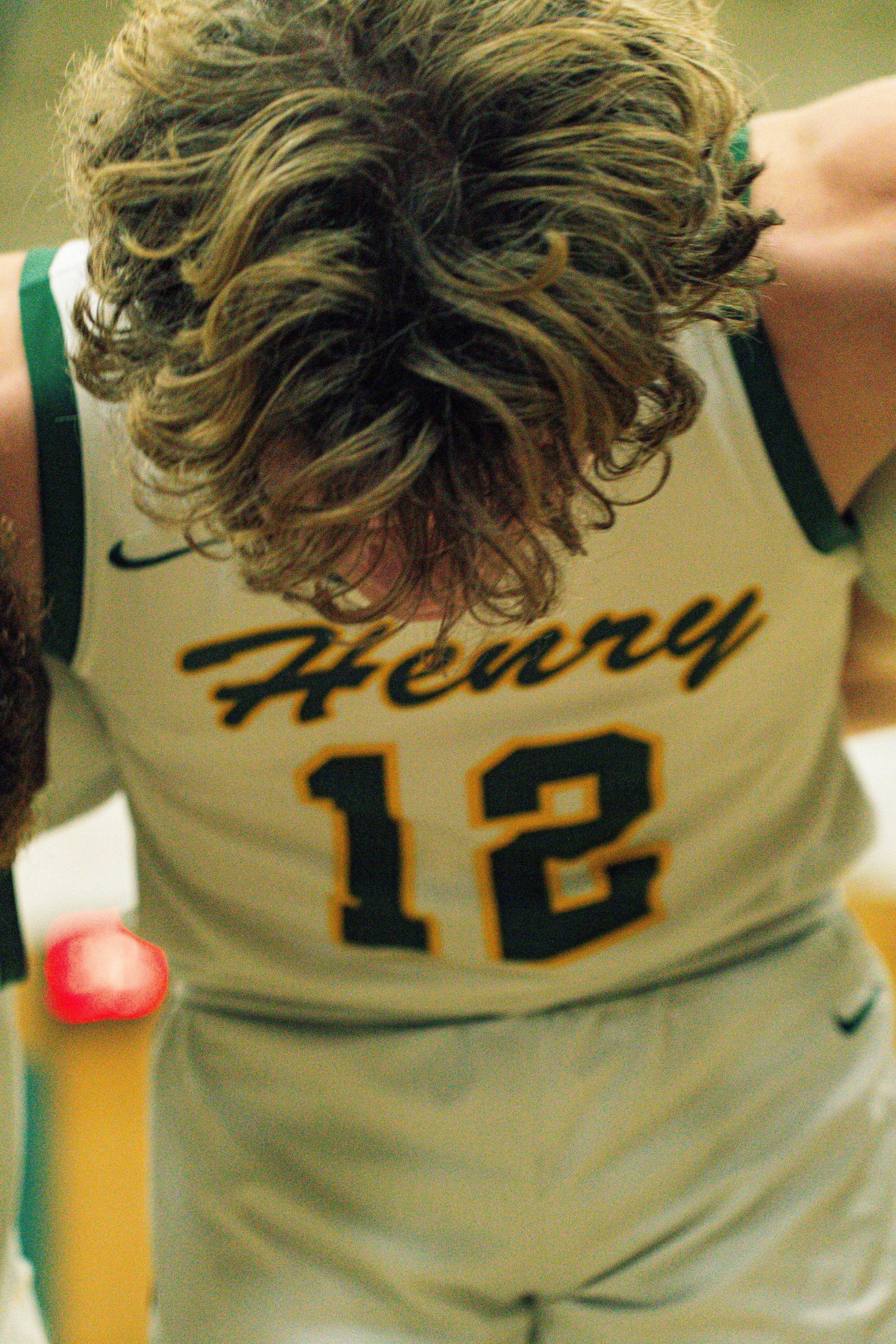 A person with long, curly hair wearing a white sports jersey with black and orange lettering and the number 13 on it, sitting at a table with some pink and yellow objects nearby.