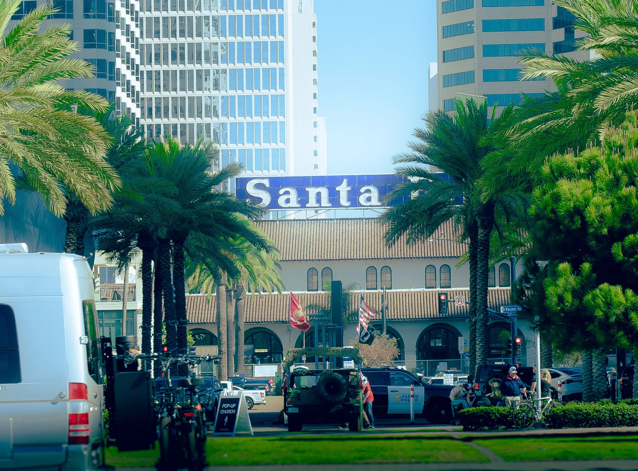 The image depicts a busy urban area with high-rise buildings, palm trees, and a parking lot filled with cars. There is a sign that reads 'Santa' on a building in the background, along with some smaller signs and flags near the parked vehicles.