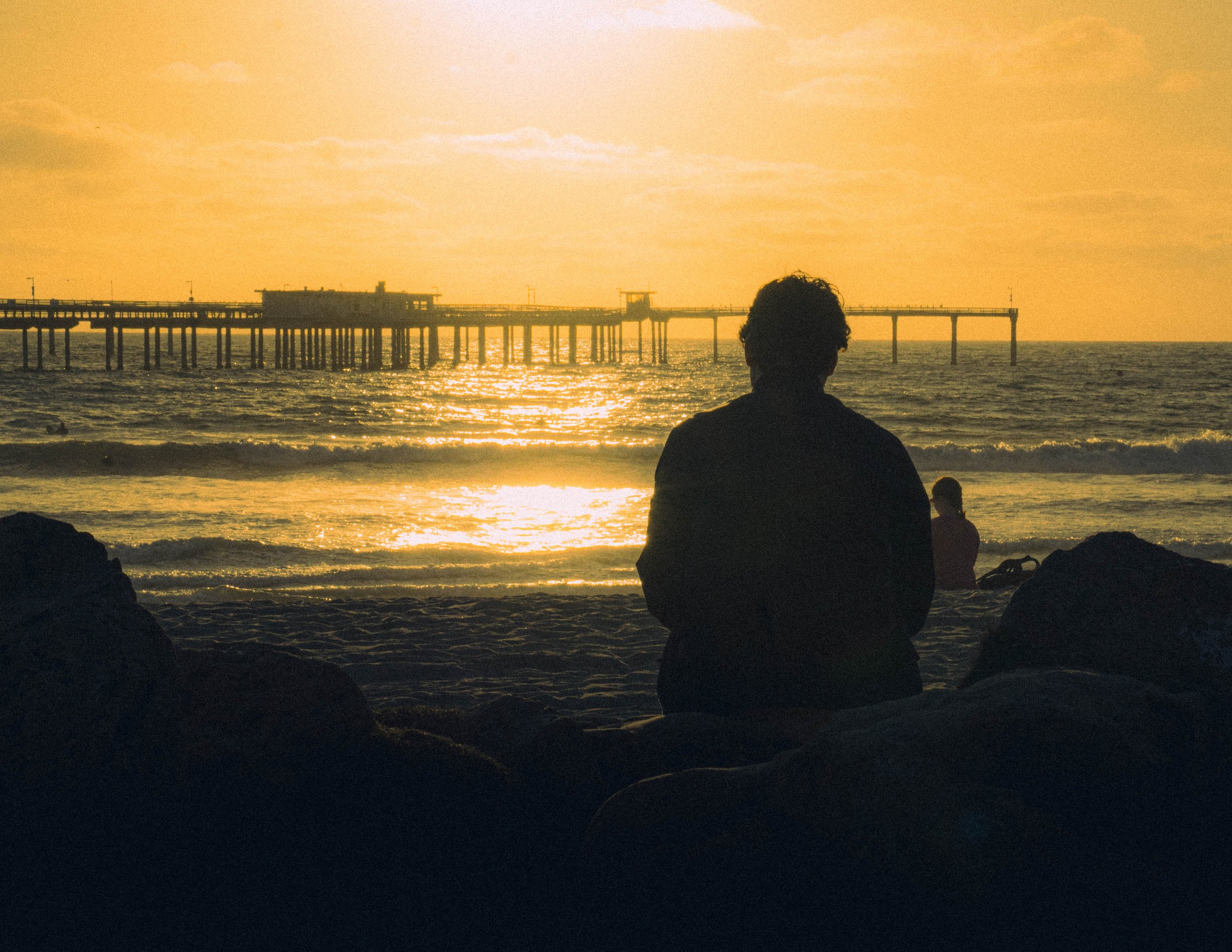 A person sitting on rocks at the beach during sunset, facing a pier extending into the ocean with the sun setting behind it.