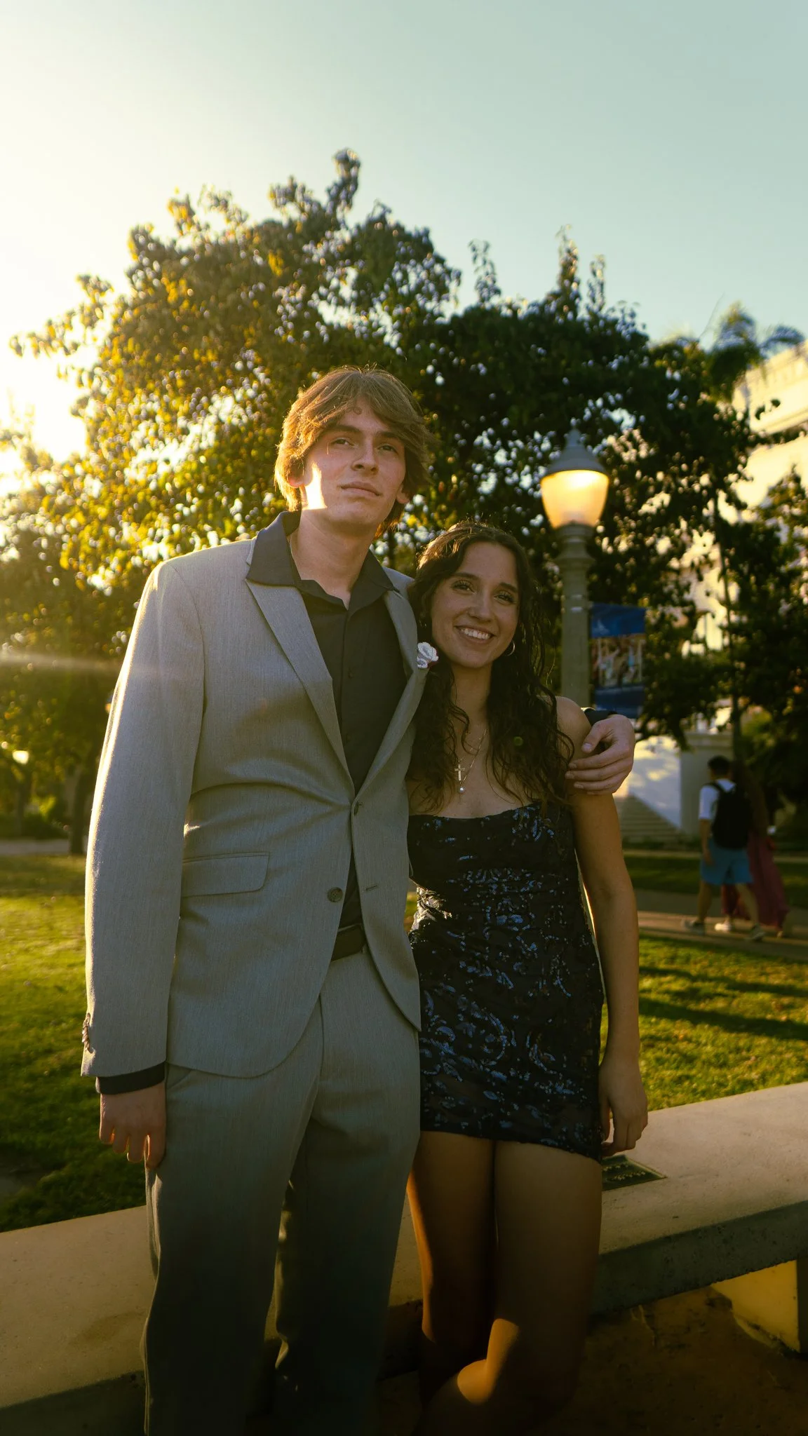A young man and woman standing together outdoors during sunset. The man is wearing a light gray suit with a black shirt, and the woman is in a black dress with floral patterns, smiling. There are trees, a lamp post, and a few people in the background