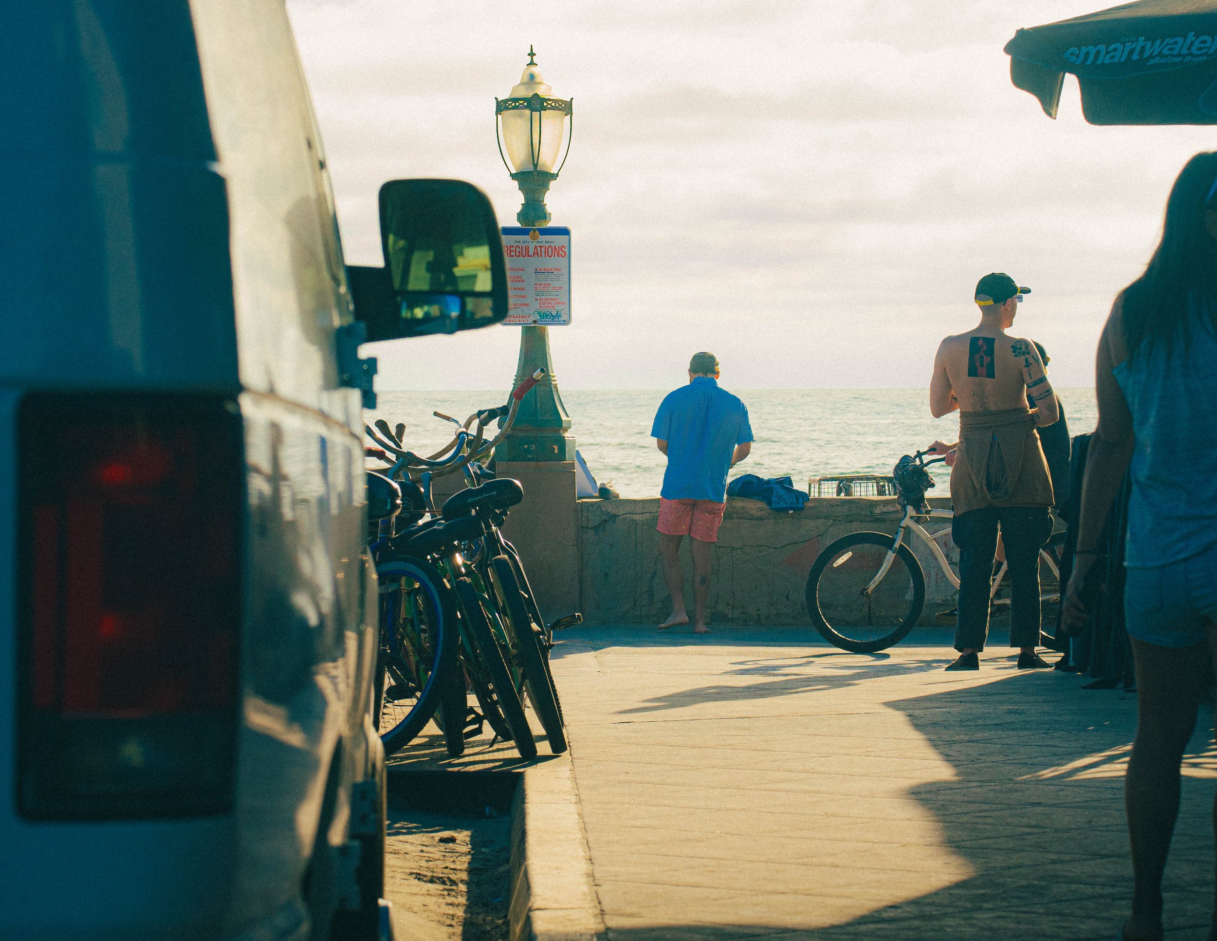 People on a walkway near the beach, with bicycles and a lamppost, under cloudy skies.
