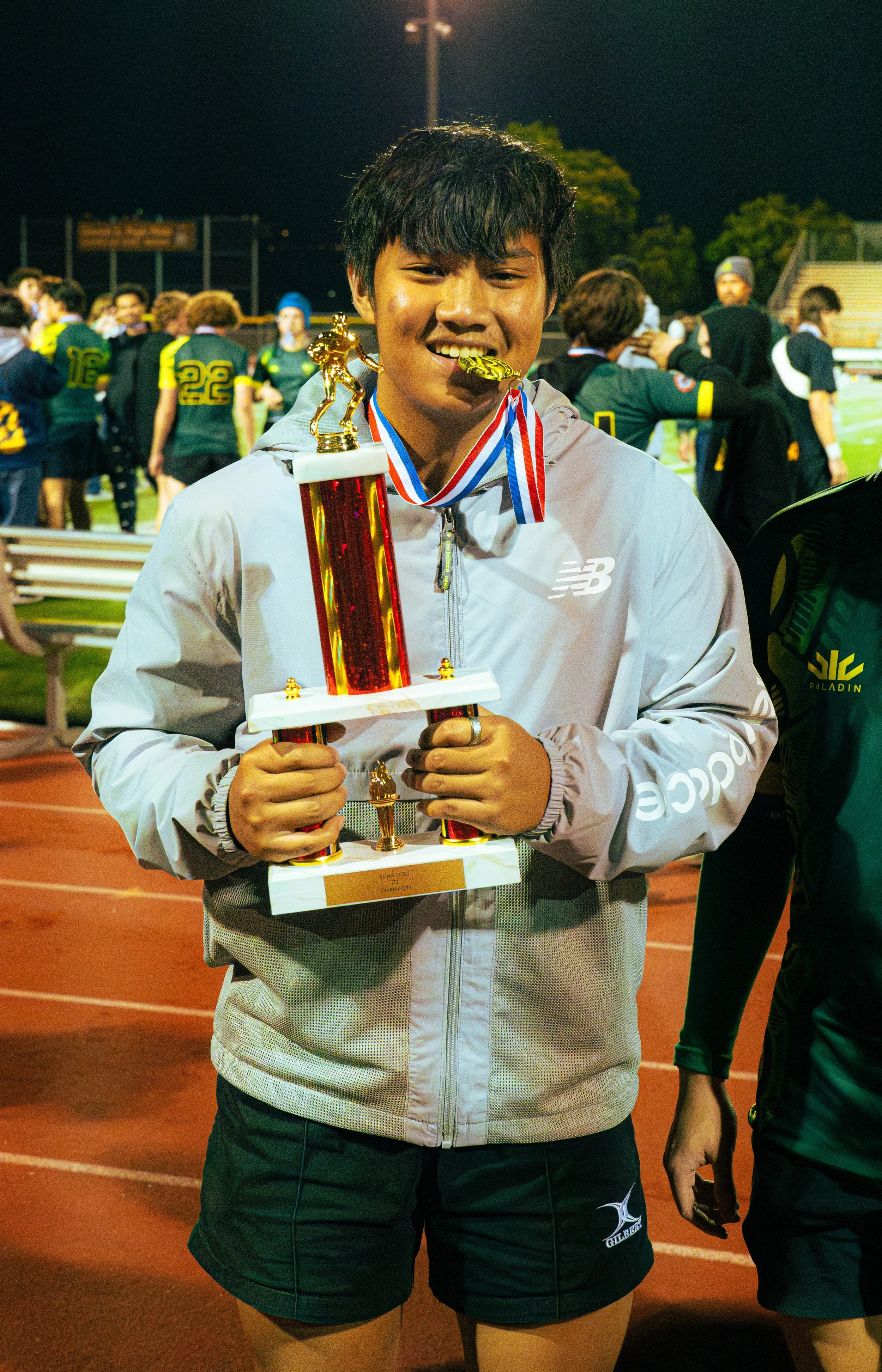 A young woman in sportswear holding a trophy with a gold football player figurine on top, smiling after a nighttime sports event, with other players and stadium seating in the background.