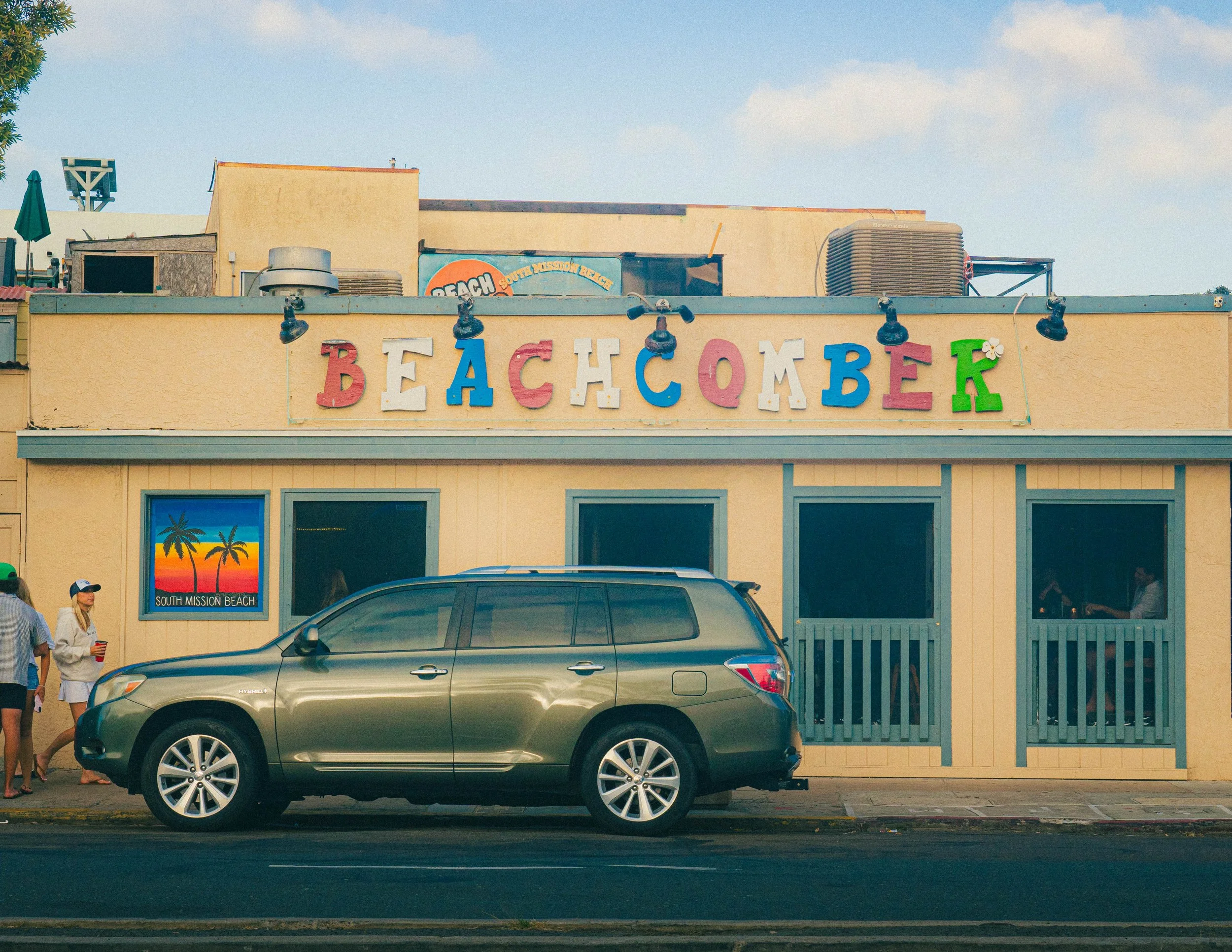 Building with colorful sign saying 'Beach Comber' in front of a blue sky, with a dark gray car parked in front and people walking nearby.