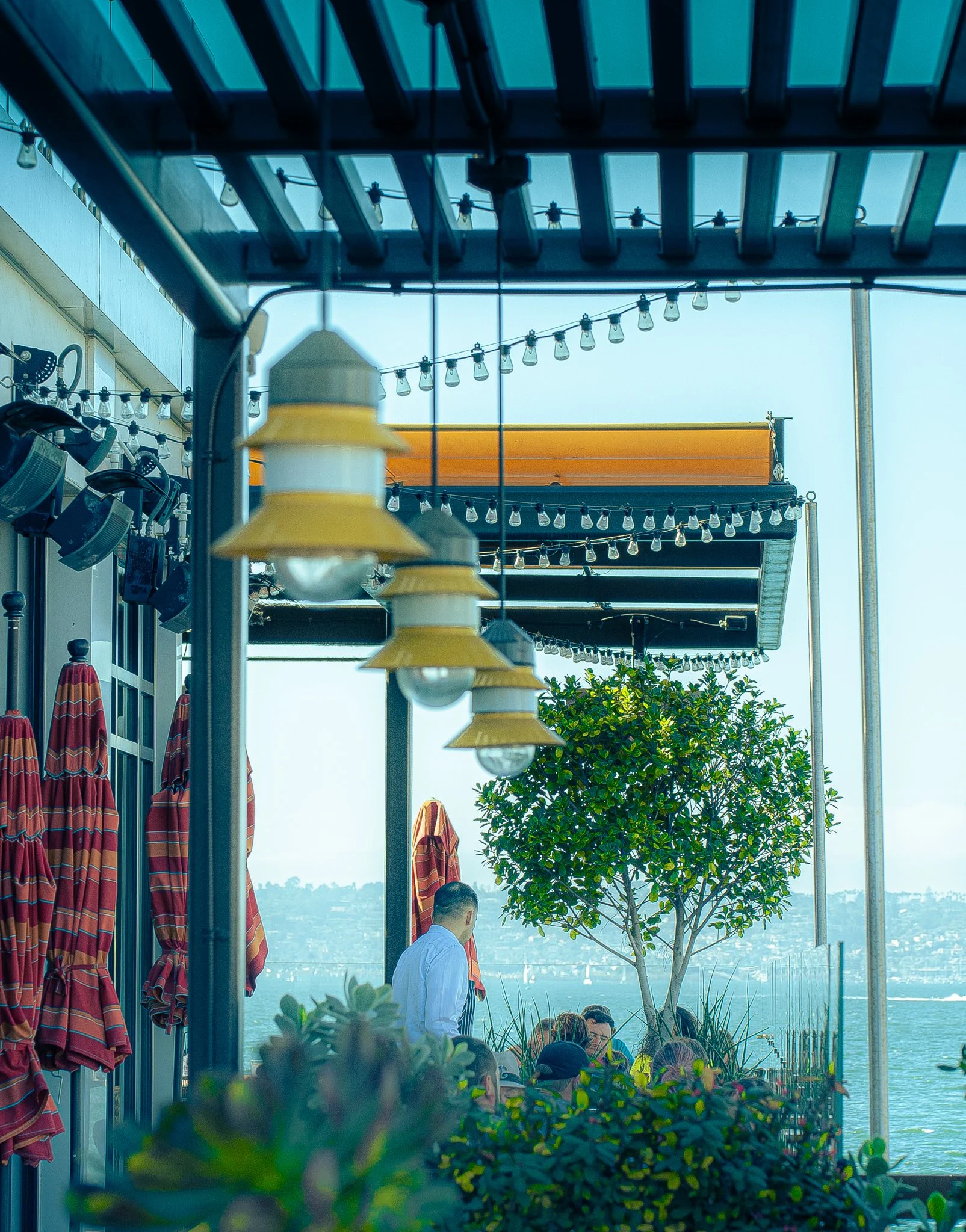 People sitting at an outdoor patio with string lights, umbrellas, and potted plants under a blue sky, overlooking a body of water.