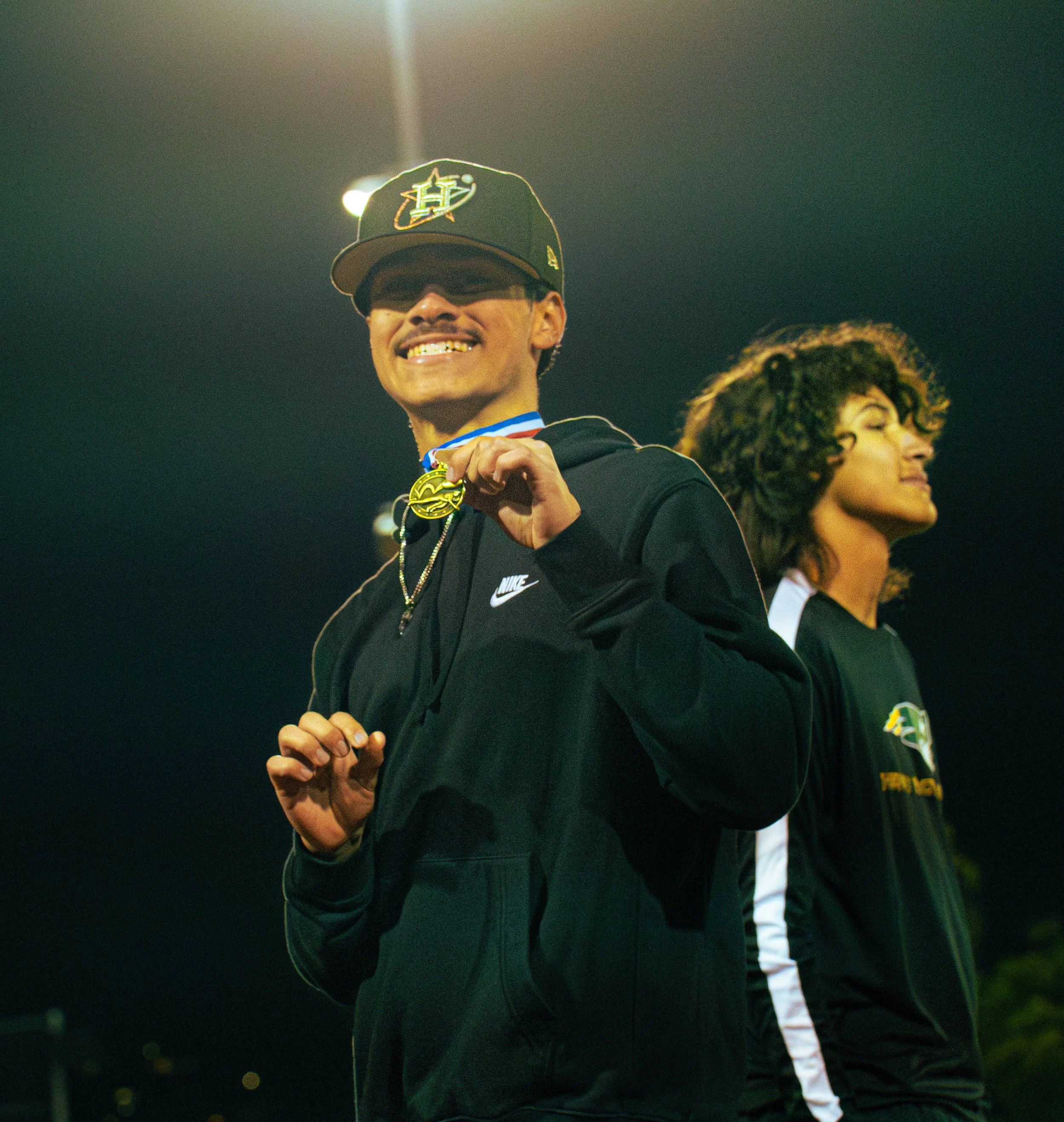 A man smiling with gold teeth, wearing a baseball cap and glasses, holding a gold medal, standing on a sports field with others in the background at night.