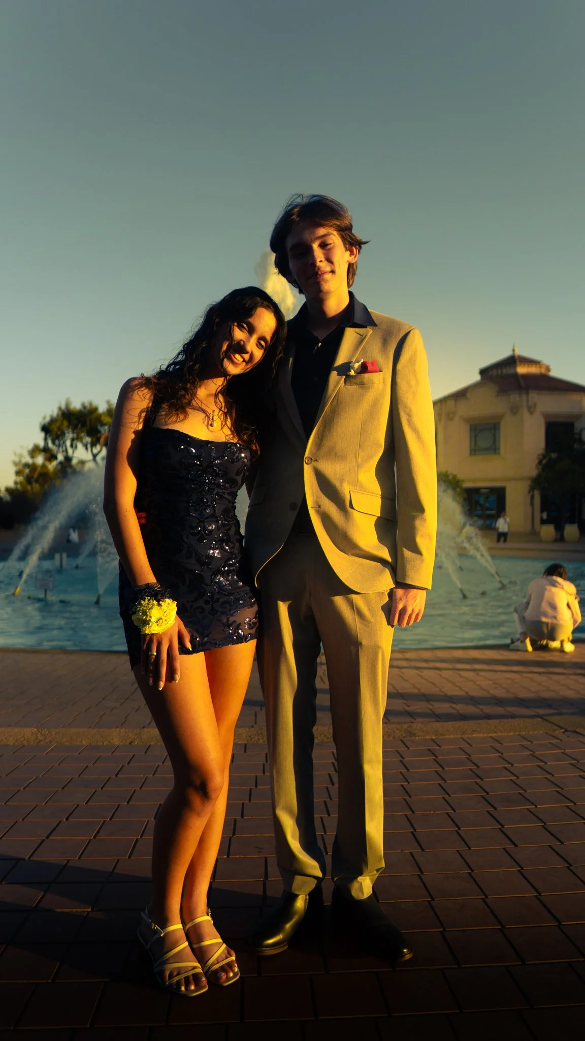 A young couple dressed in formal attire stands together near a fountain with a large building in the background during sunset.