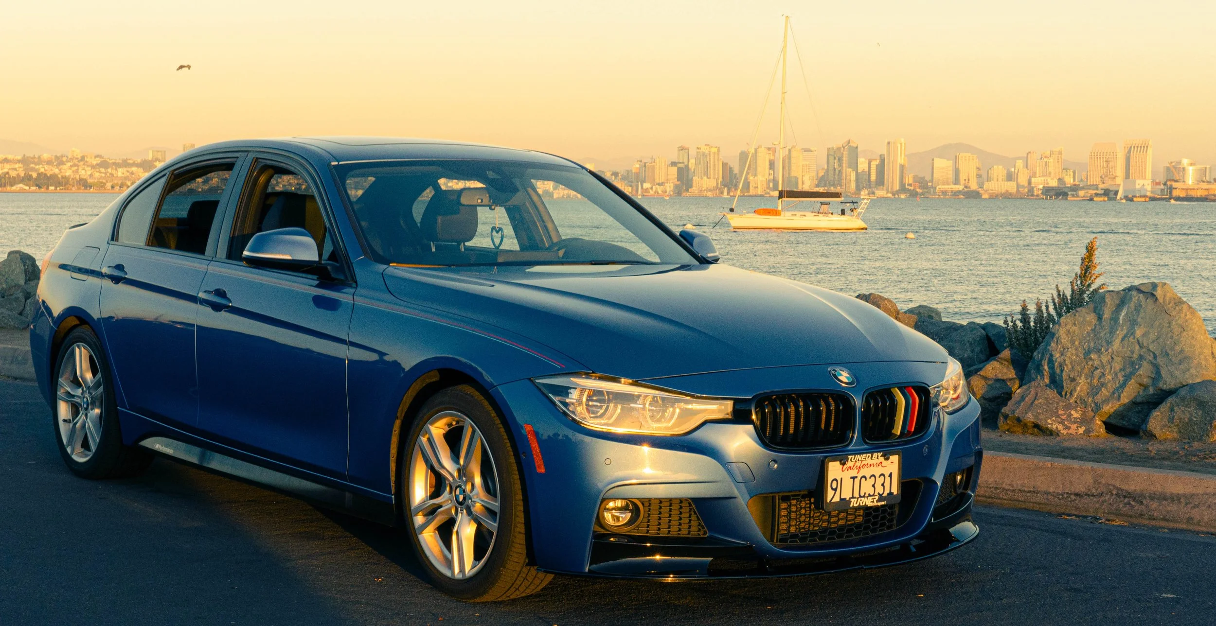 Blue BMW sedan parked near the water with a city skyline and sailboat in the background during sunset.