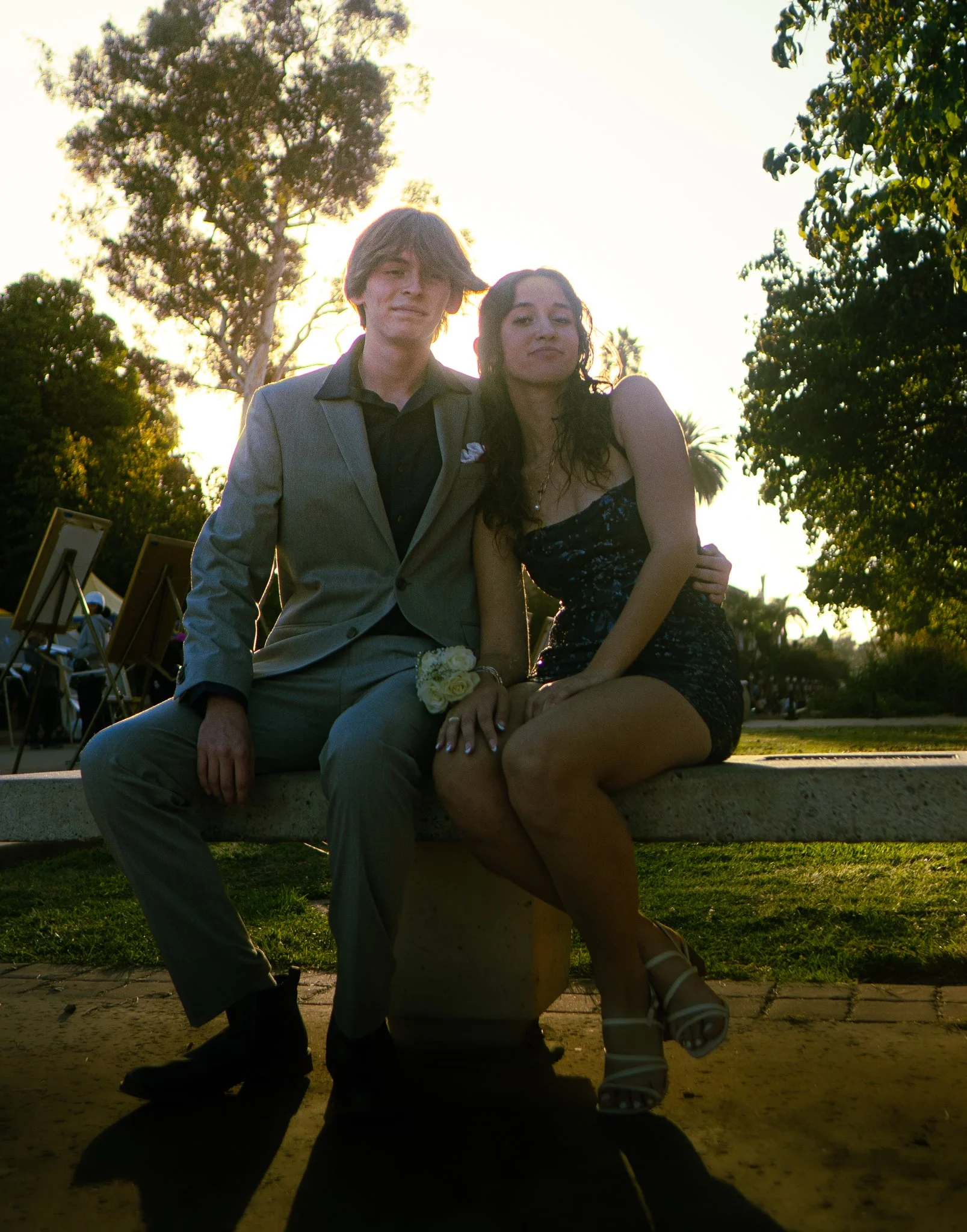 A young man and woman sitting on a park bench during sunset, with trees and a bright sky in the background. The man wears a light gray suit and the woman wears a black dress with white floral patterns, white heels, and has long, curly dark hair.