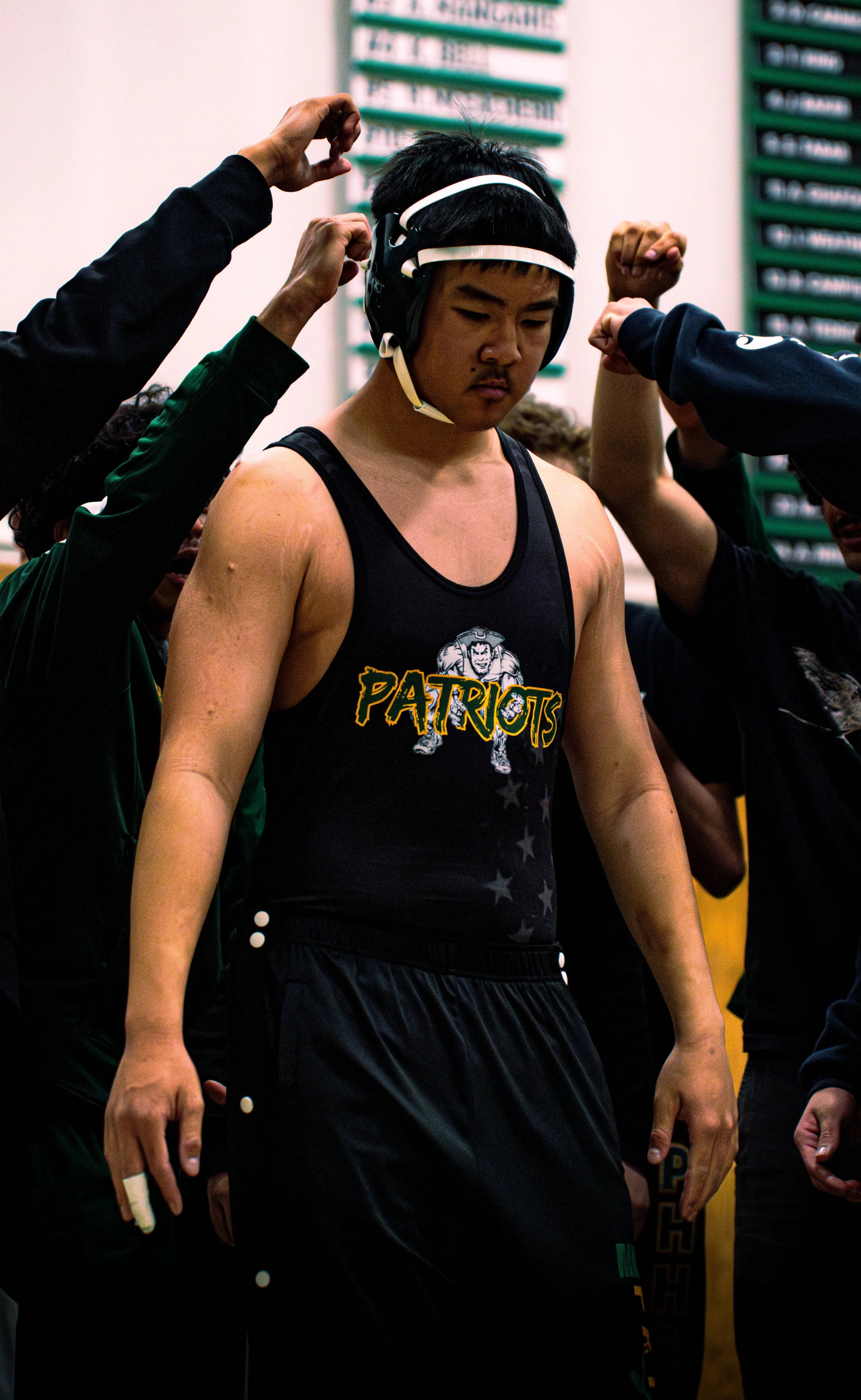 A young male wrestler wearing a black and green Patriots singlet and a black protective headgear stands with his head bowed as others adjust his headgear.