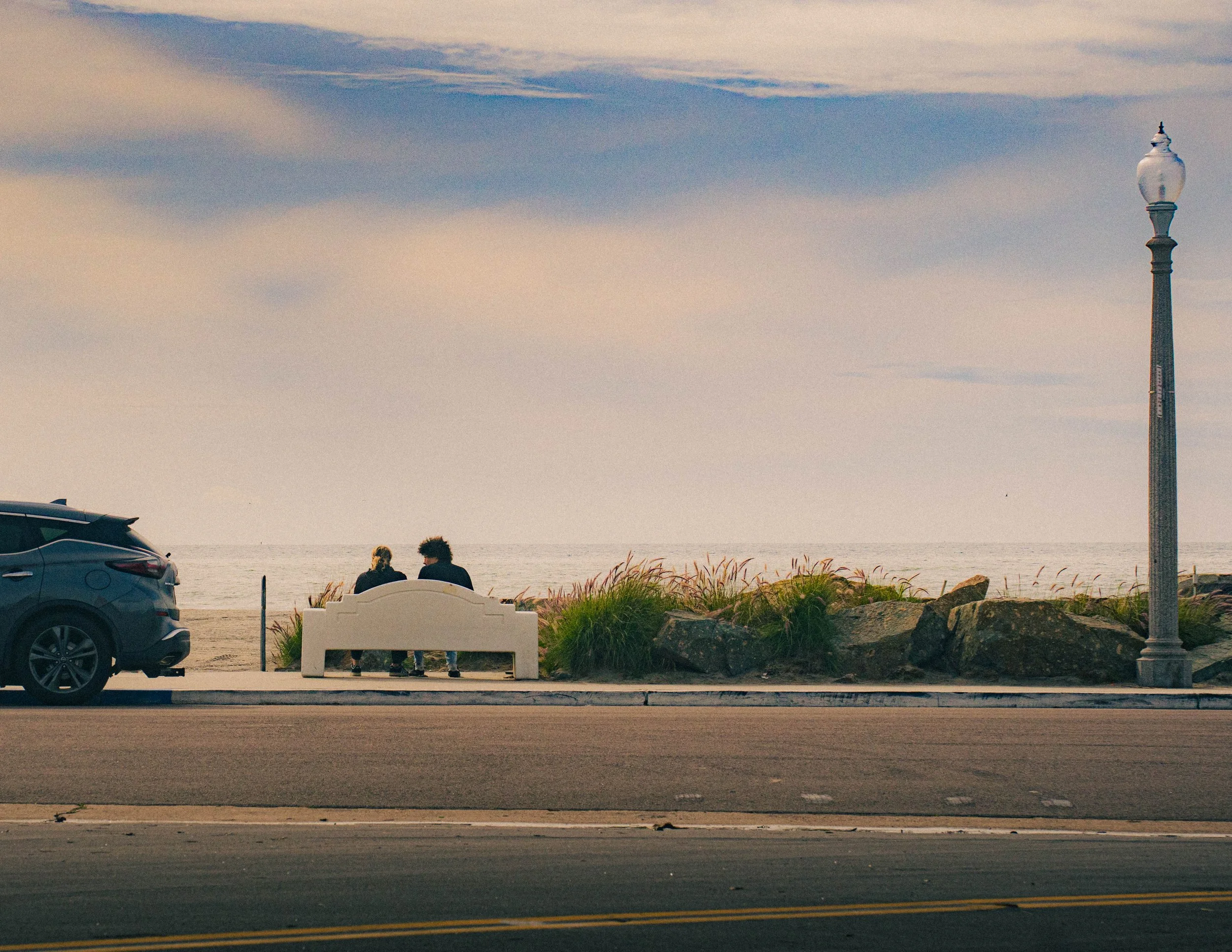 A beach scene with a street in the foreground, a car parked on the left, two people sitting on a bench facing the ocean, and a tall structure with a decorative top in the background under a sky with clouds.