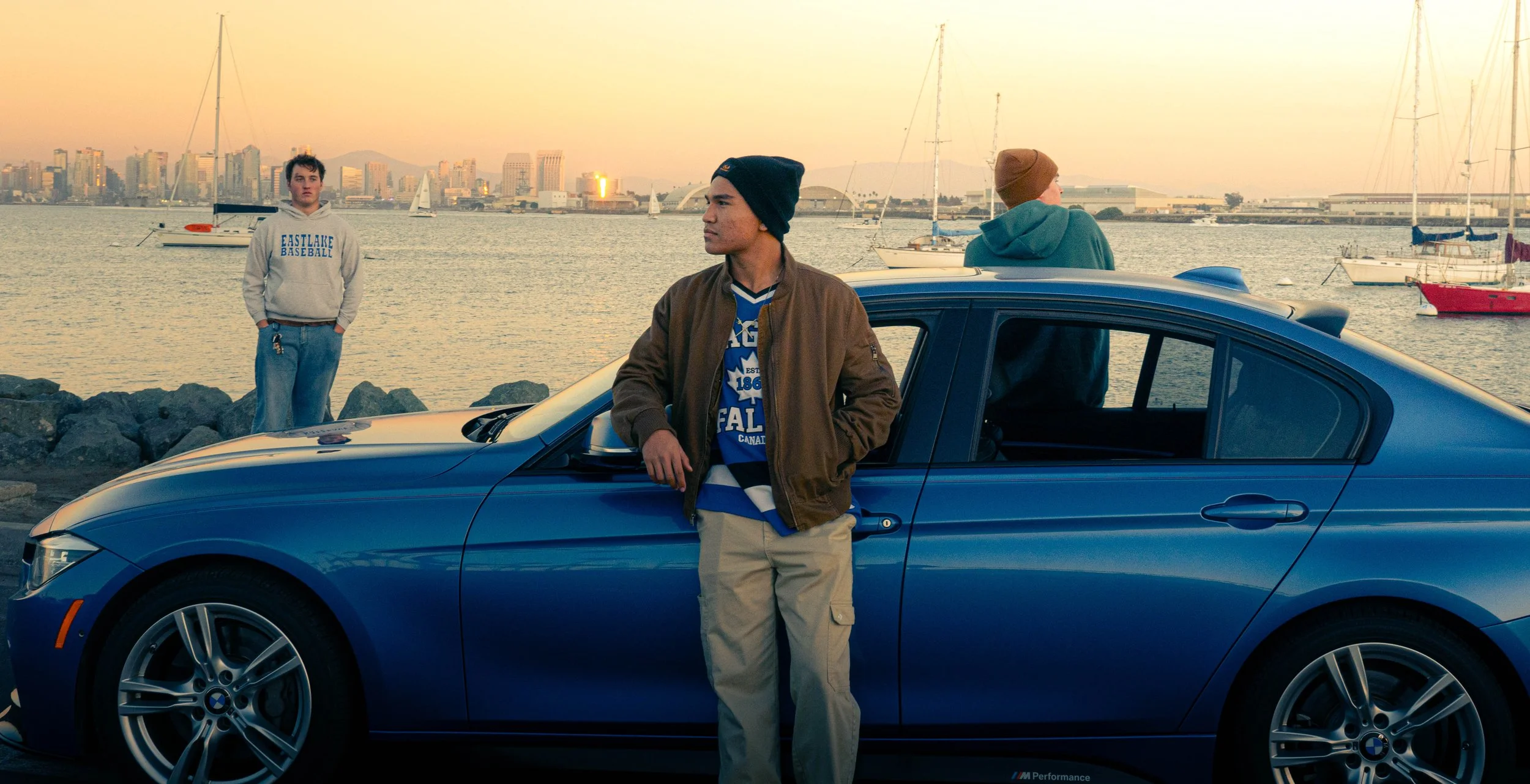 Three young men by a blue BMW car parked near a waterfront at sunset, with city skyline and sailboats in the background.