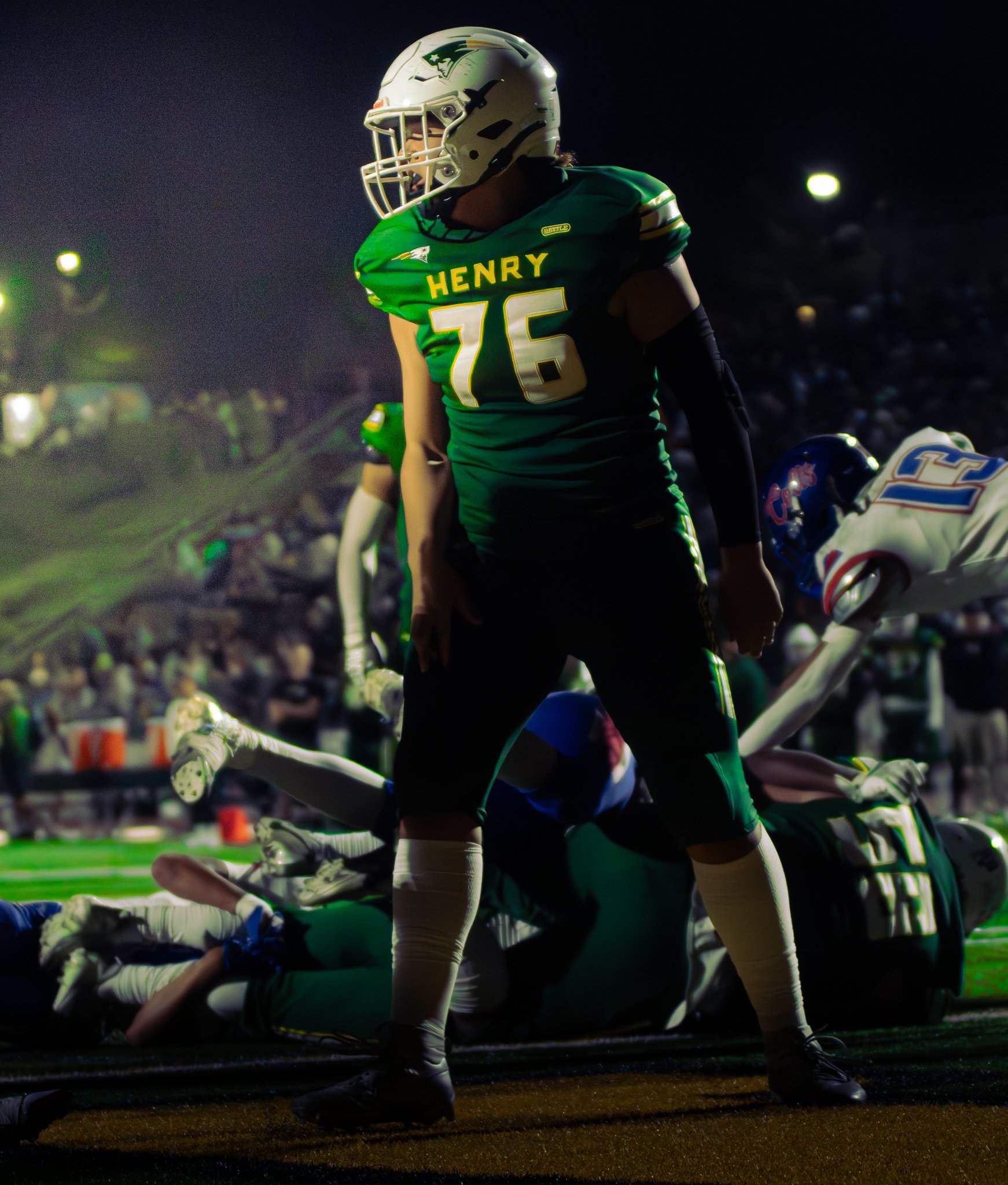 A football player from Henry High School, number 76, wearing a green uniform and white helmet, is kneeling during a game at night. Other players and spectators are visible in the background.