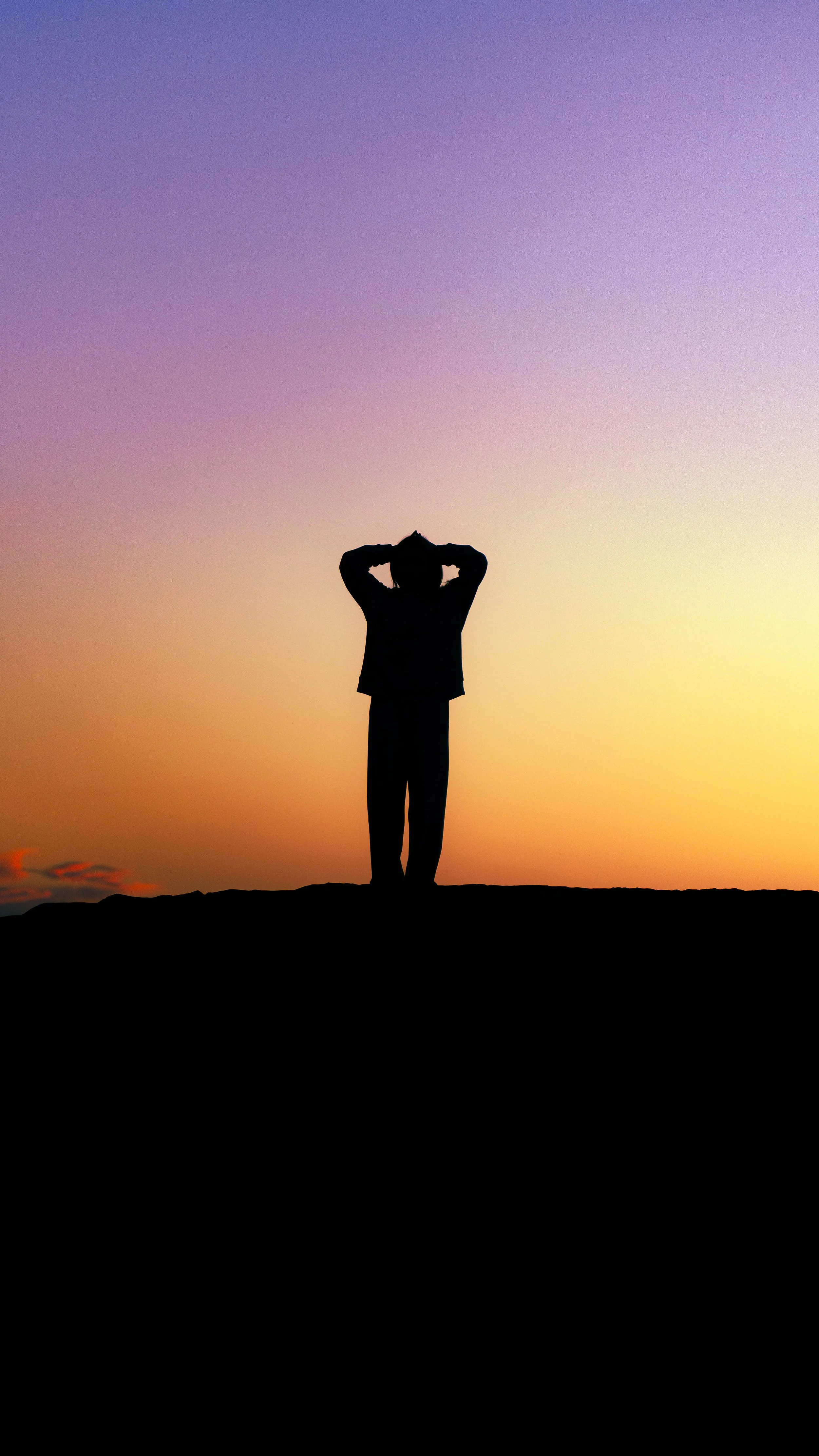 Silhouette of a person standing on a horizon at sunset, with arms raised behind their head, against a colorful sky with shades of purple, pink, and yellow.
