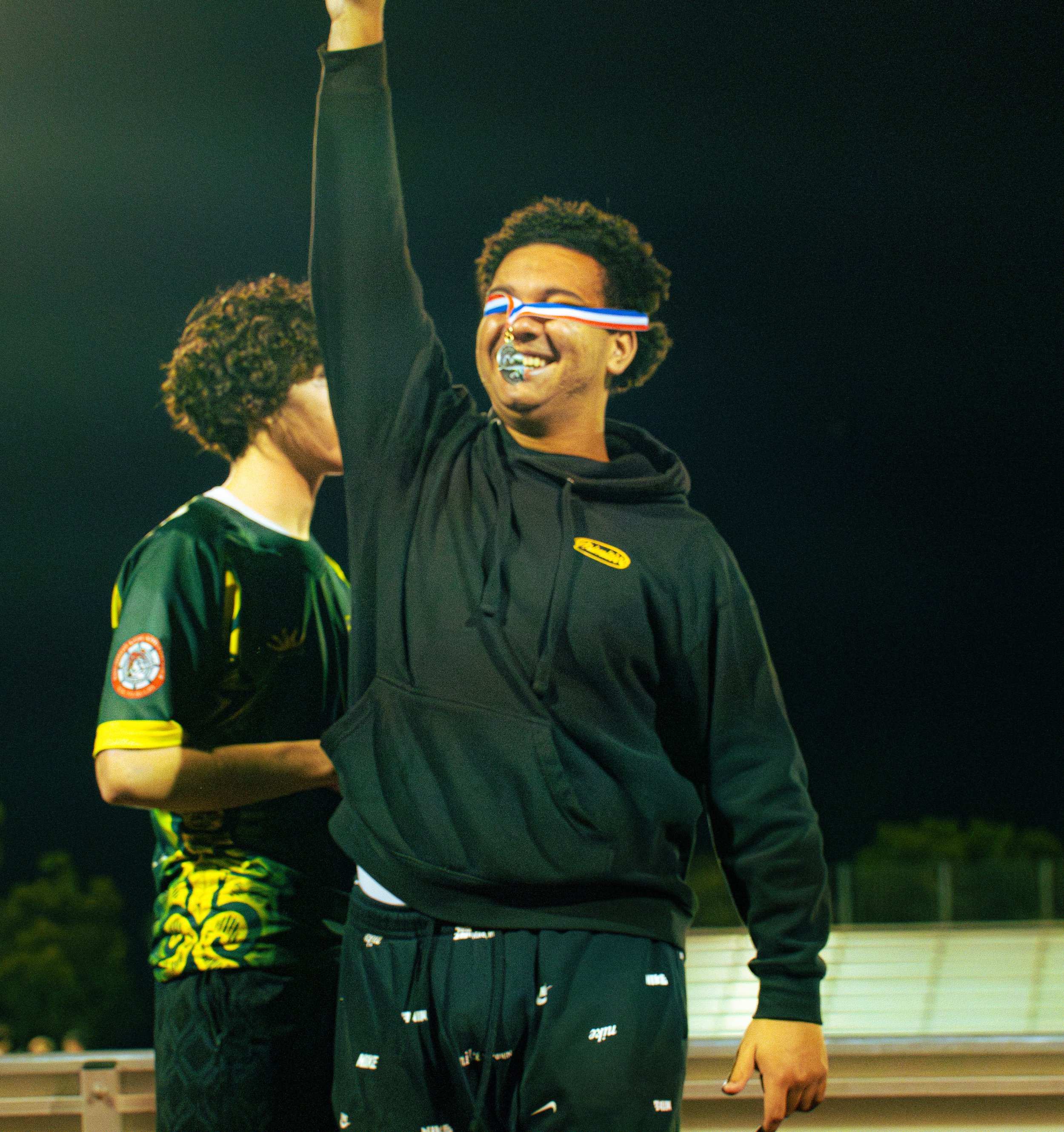 Two young men interact indoors at a social event; one smiling with a medal around his neck, wearing a headband with red, white, and blue stripes, and a black hoodie, the other with curly hair, wearing a patterned shirt and shorts.