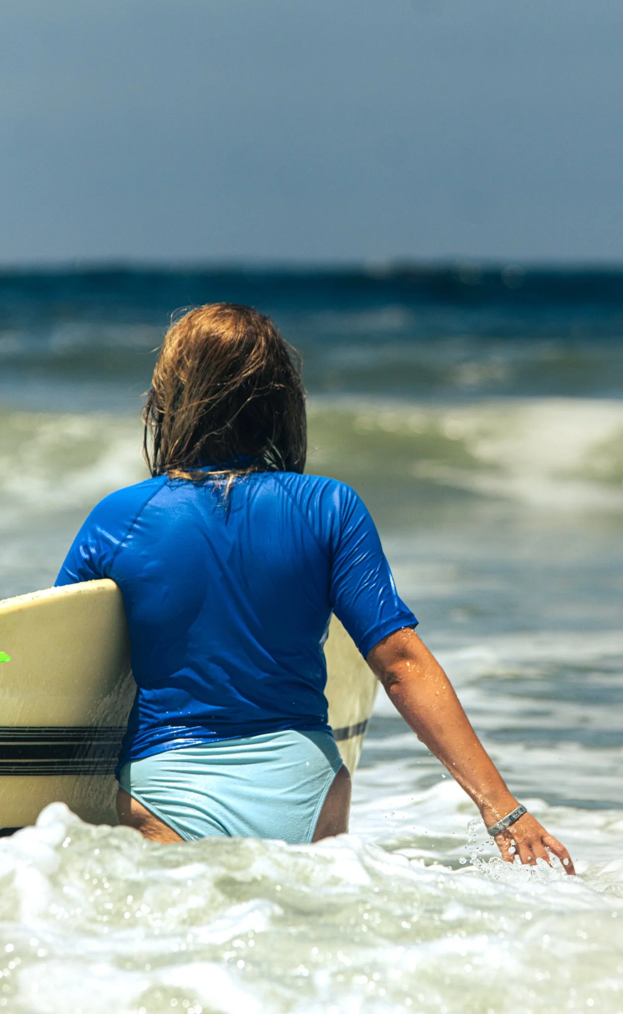 A person with long hair wearing a blue shirt and light blue shorts sitting on the edge of a surfboard in the ocean with waves in the background.