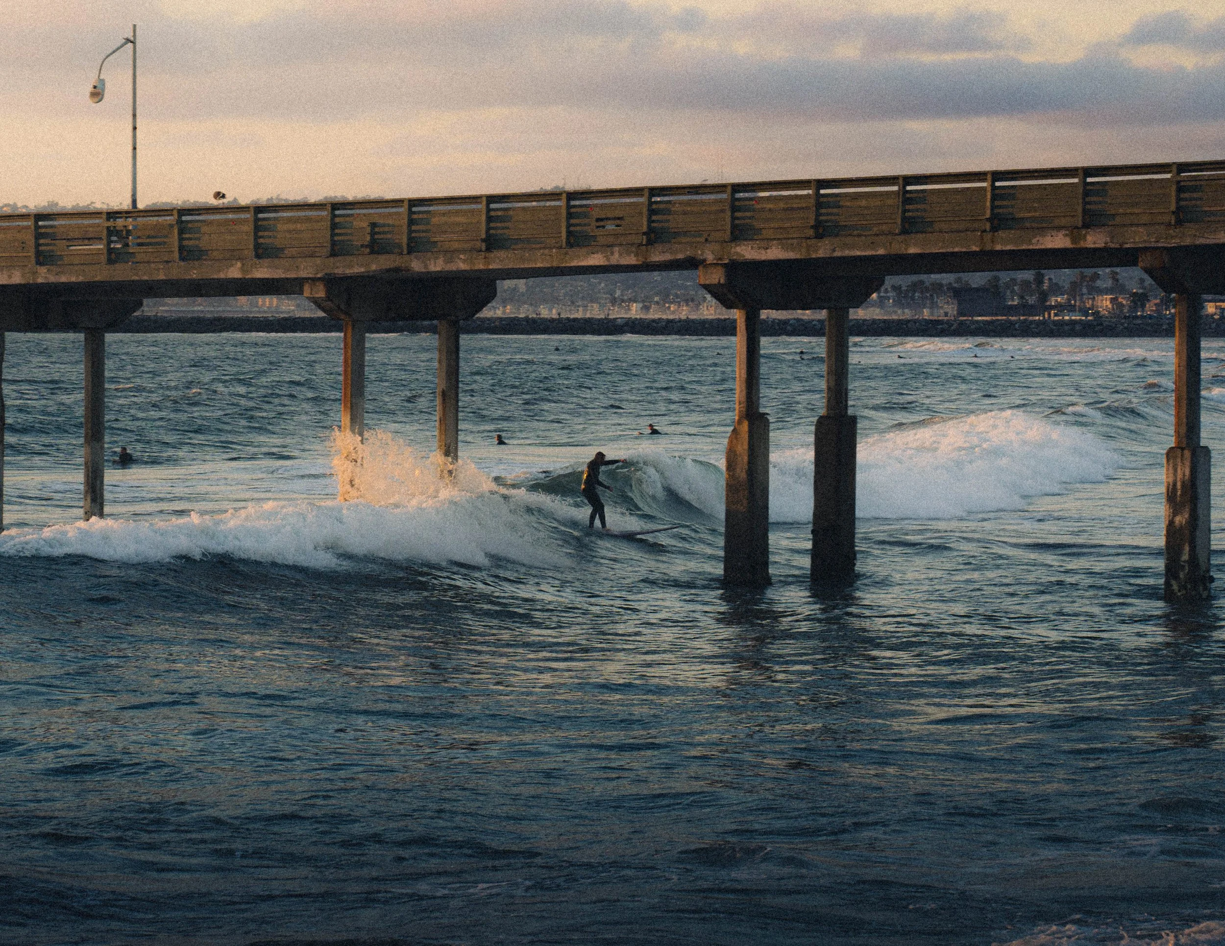 Surfers riding waves near a pier at sunset, with wooden pilings supporting the pier and city buildings in the background.