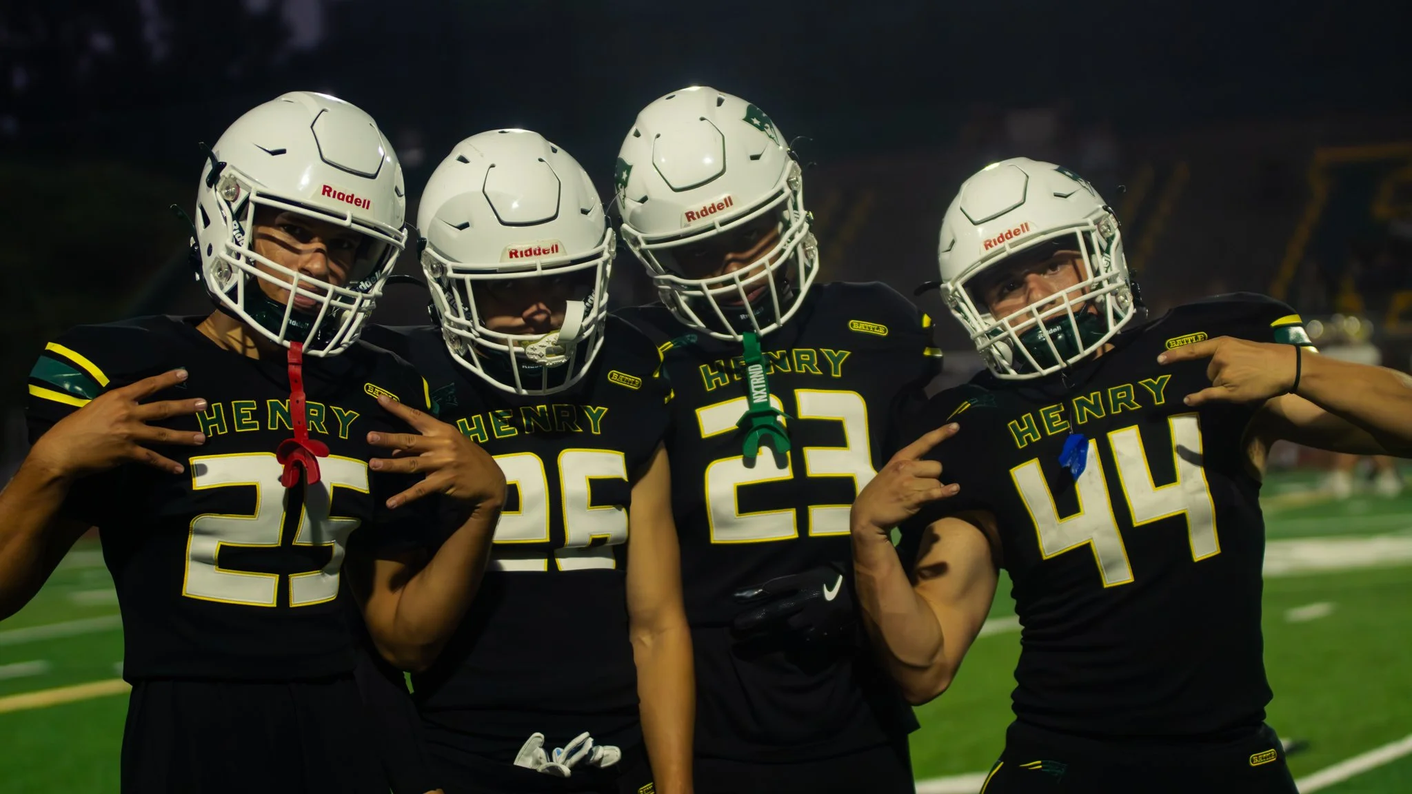Four football players wearing black jerseys with yellow and green accents and white helmets standing on a football field at night. They are posing with smiles and hand gestures.