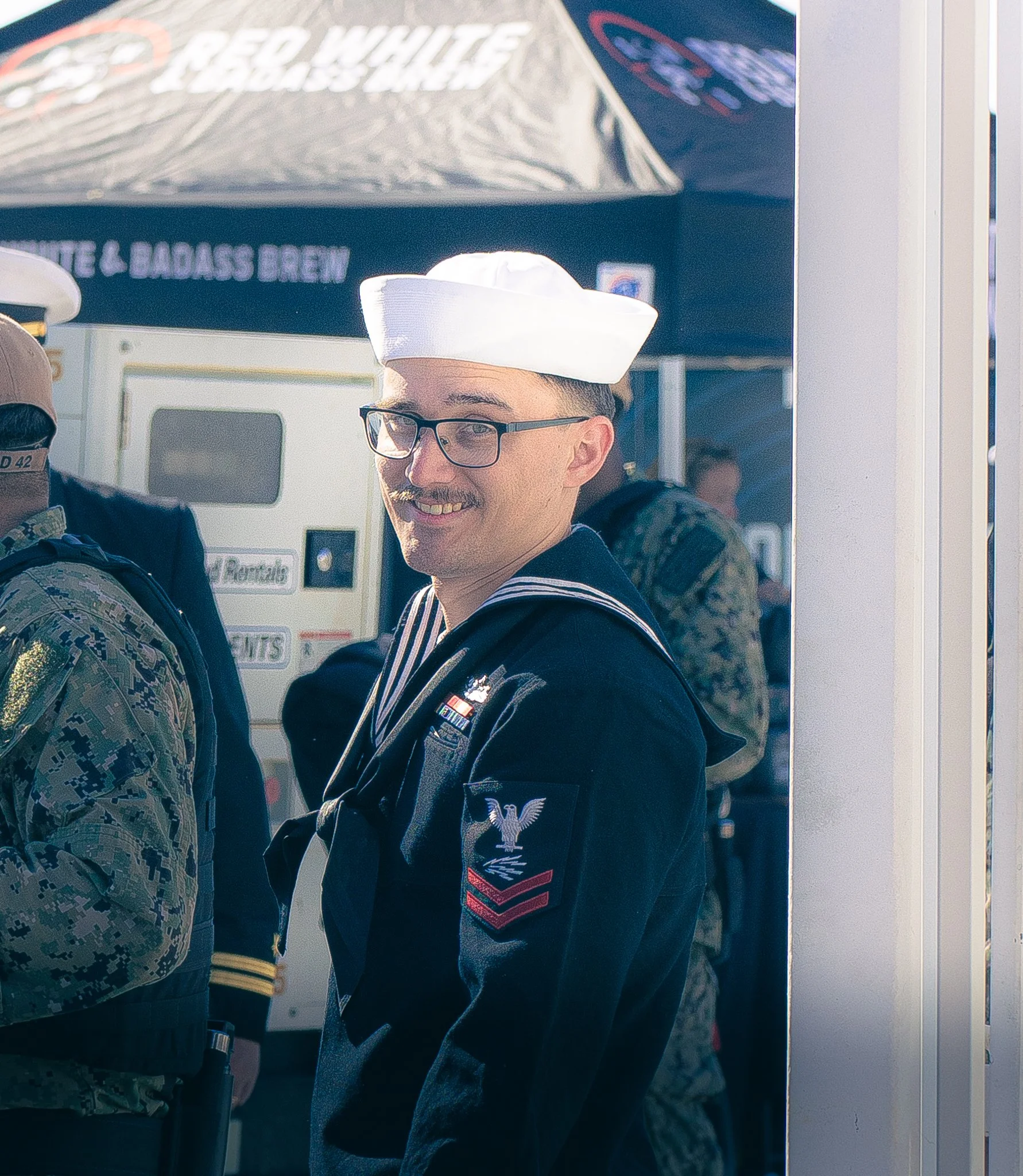 A young man wearing glasses and a Navy dress uniform with a military insignia, standing outdoors with a man in camouflage uniform and a Marine Corps cap nearby. There are structures and signs in the background.