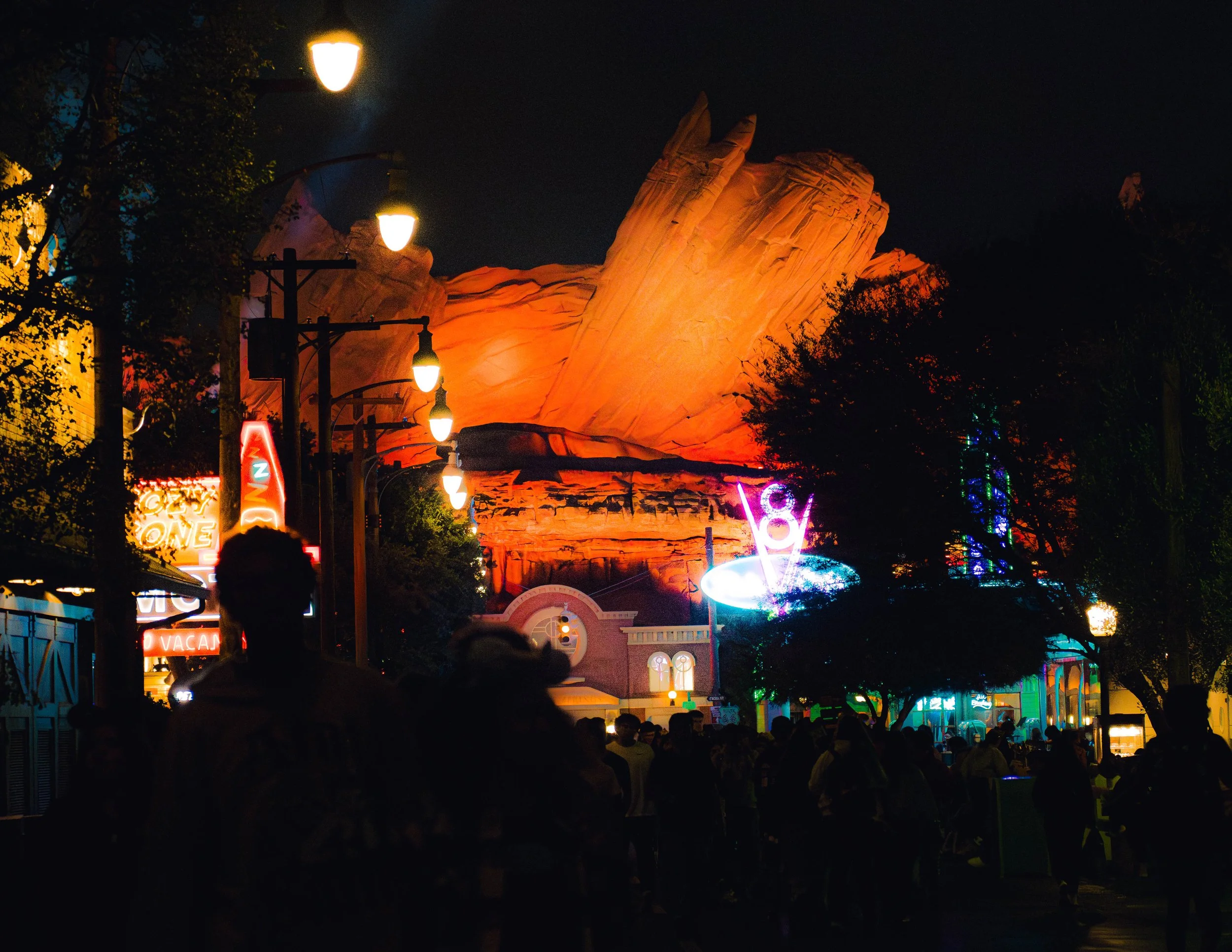 Night scene at a theme park with large illuminated rock formations in the background. Streetlights and colorful neon signs light up the area, with a crowd of people walking and standing in the foreground.