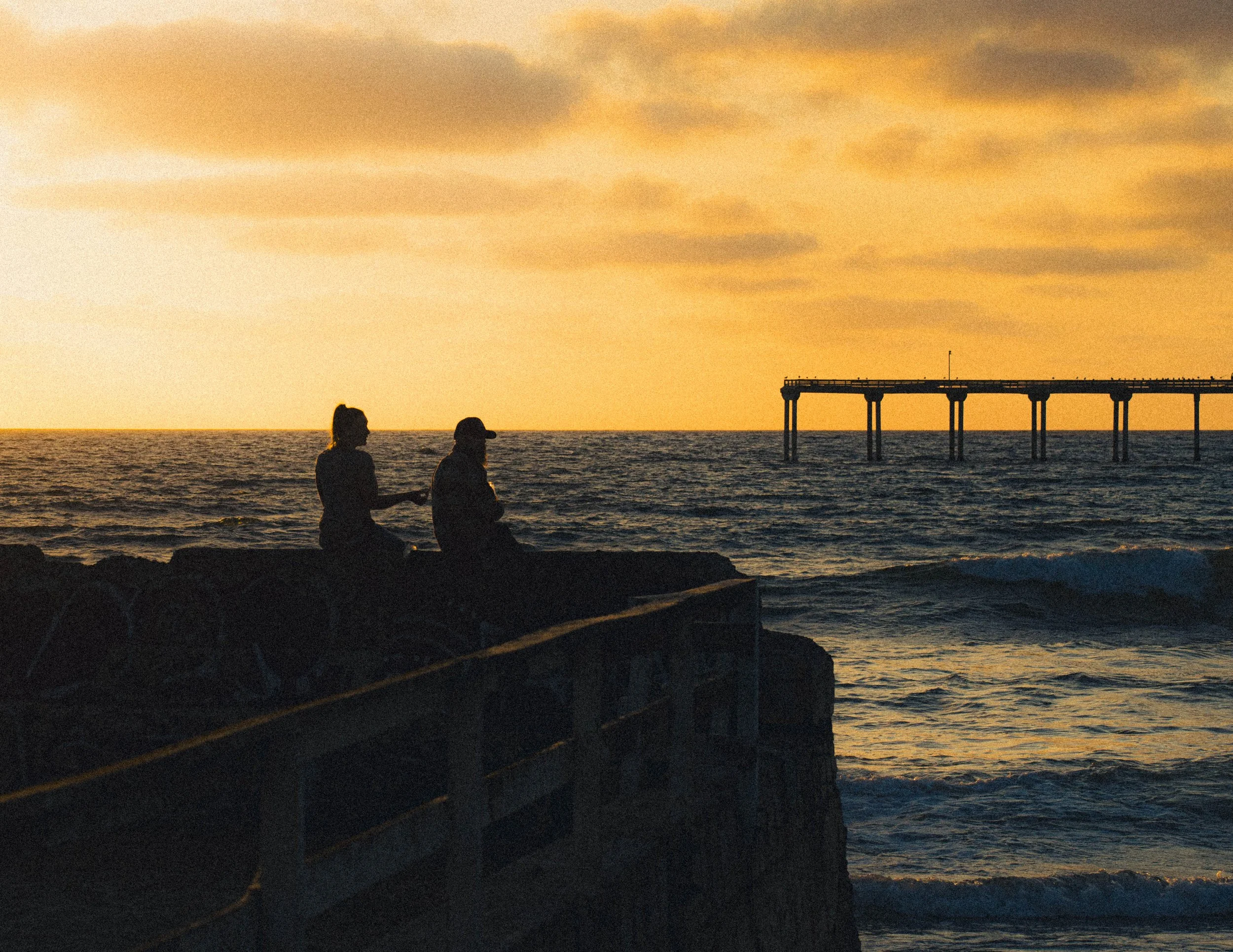 People sitting on rocks by the ocean at sunset with a pier extending into the water and cloudy sky.
