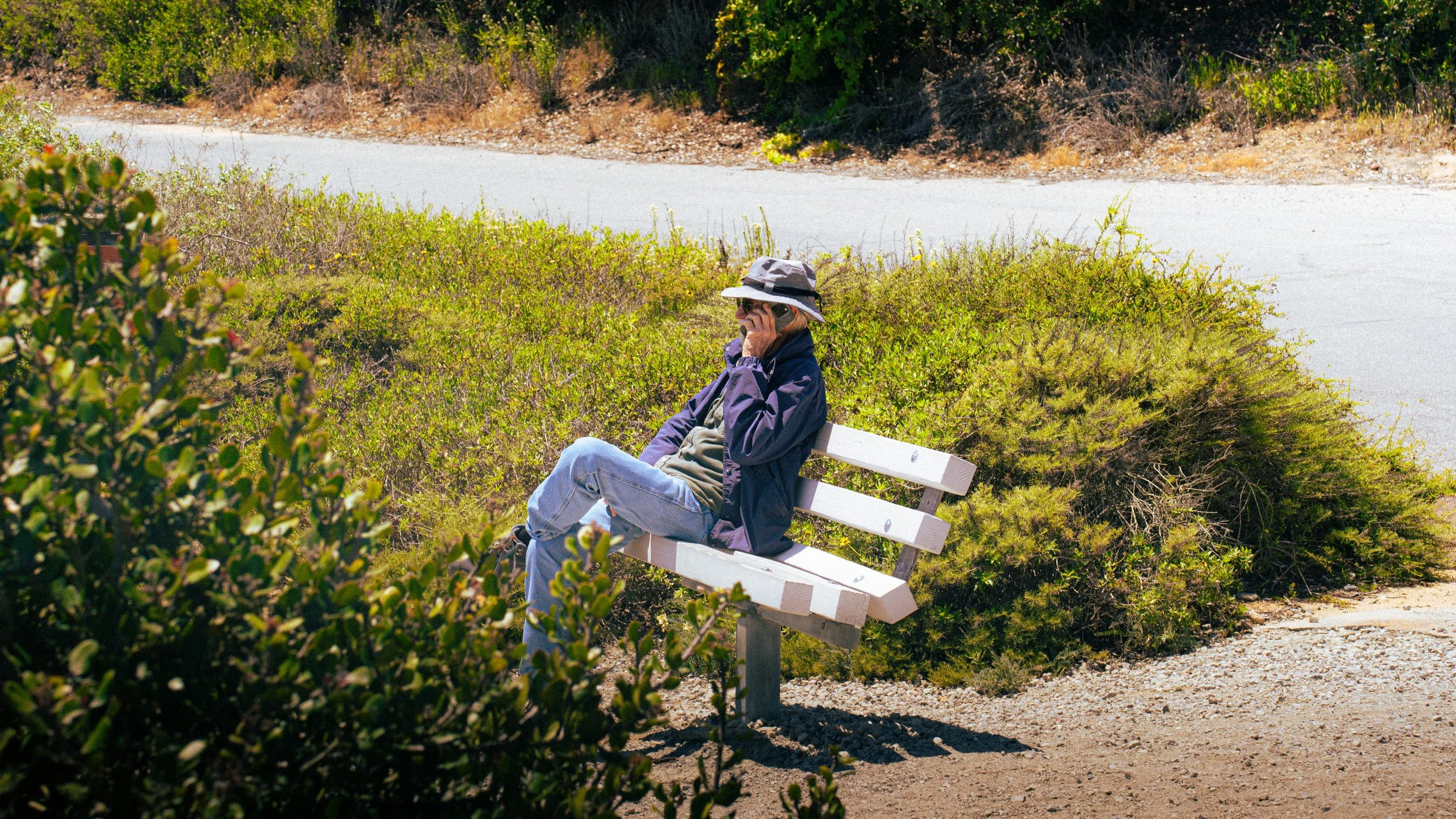 A person sitting on a white park bench in a natural outdoor setting, talking on a cellphone. The person is wearing a hat, sunglasses, a jacket, and jeans. The background features lush green bushes and a dirt pathway.