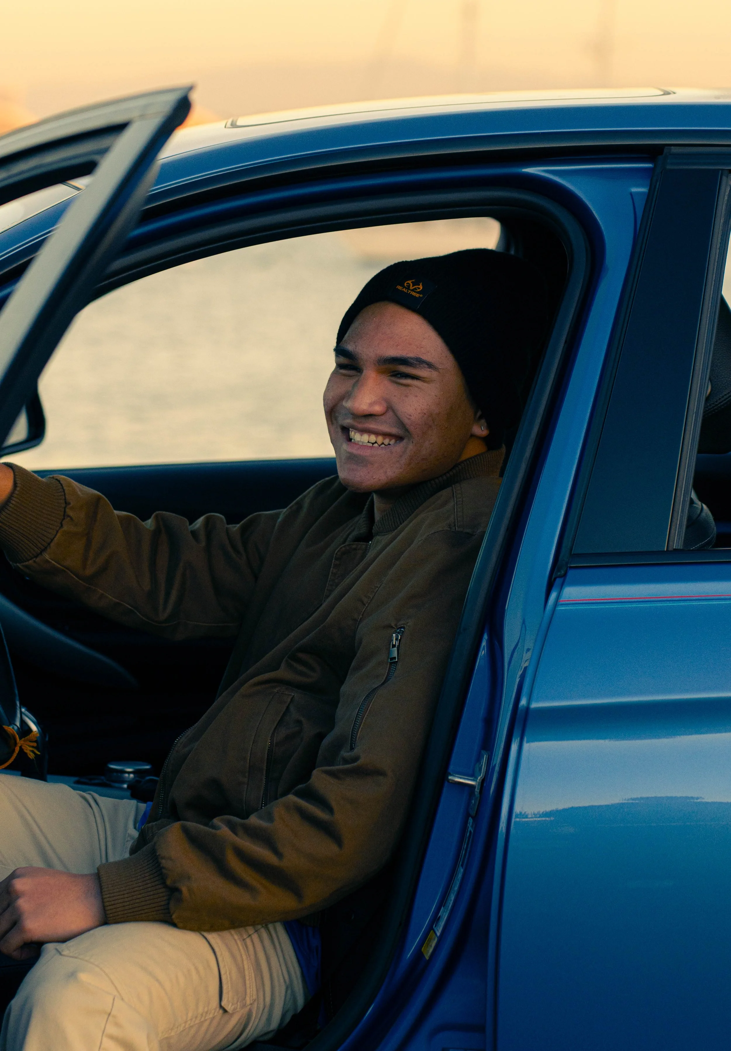 Young man driving a blue car, smiling, wearing a black beanie and brown jacket.