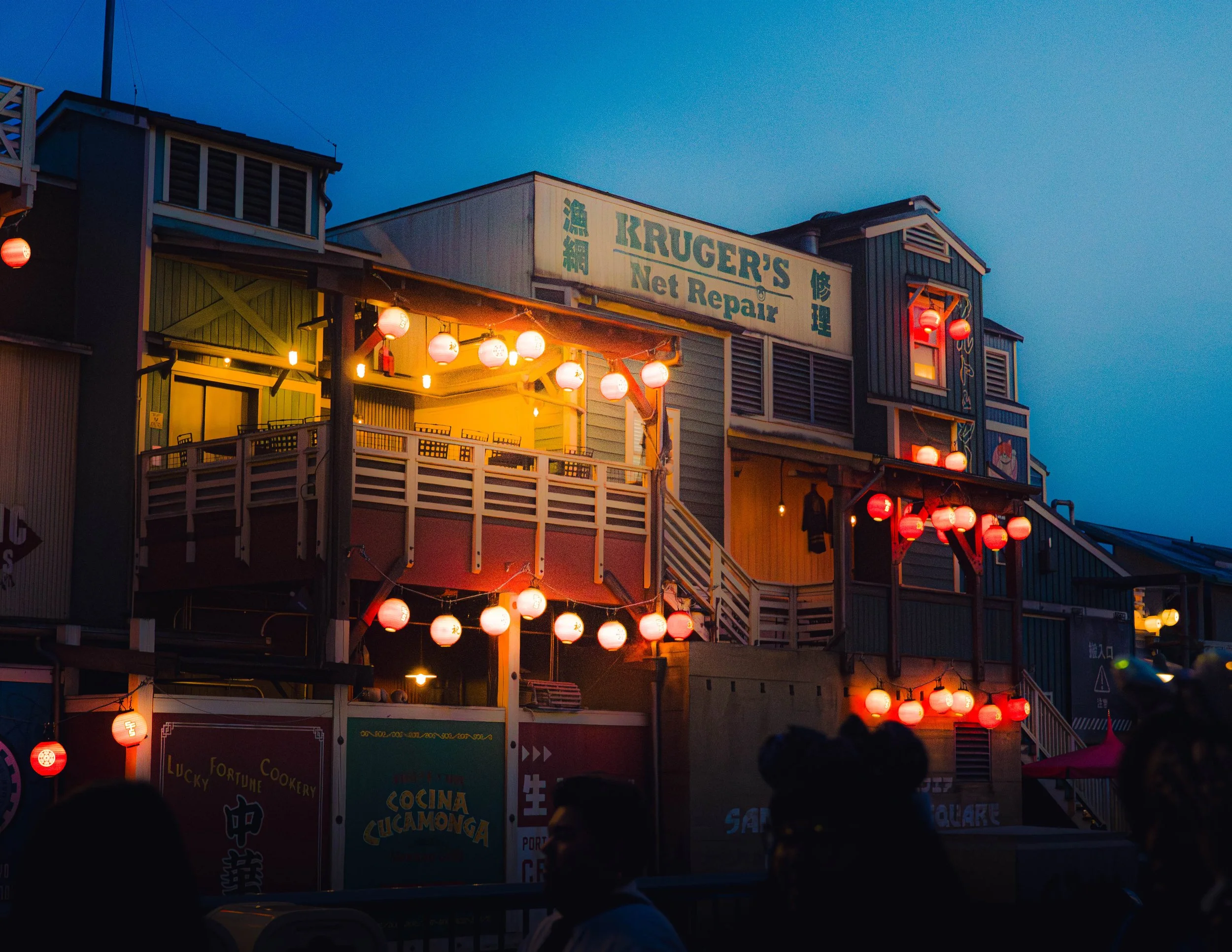 Street scene at dusk with buildings decorated with red paper lanterns and a sign for Kruger’s Net Repair, with other signs in multiple languages, and people in the foreground.