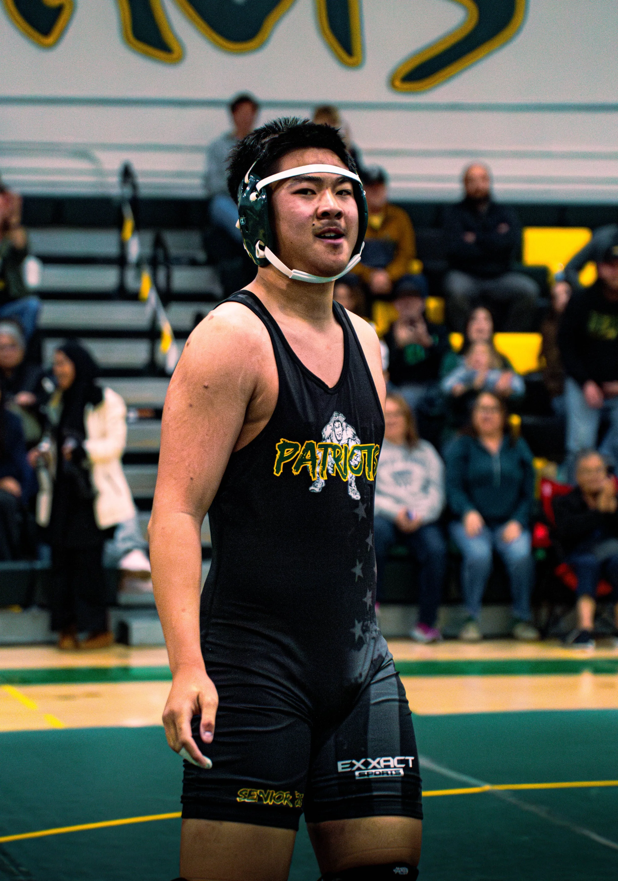 A male athlete in a black sports jersey and shorts, wearing a protective headgear, standing on a gymnasium floor with spectators in the background.