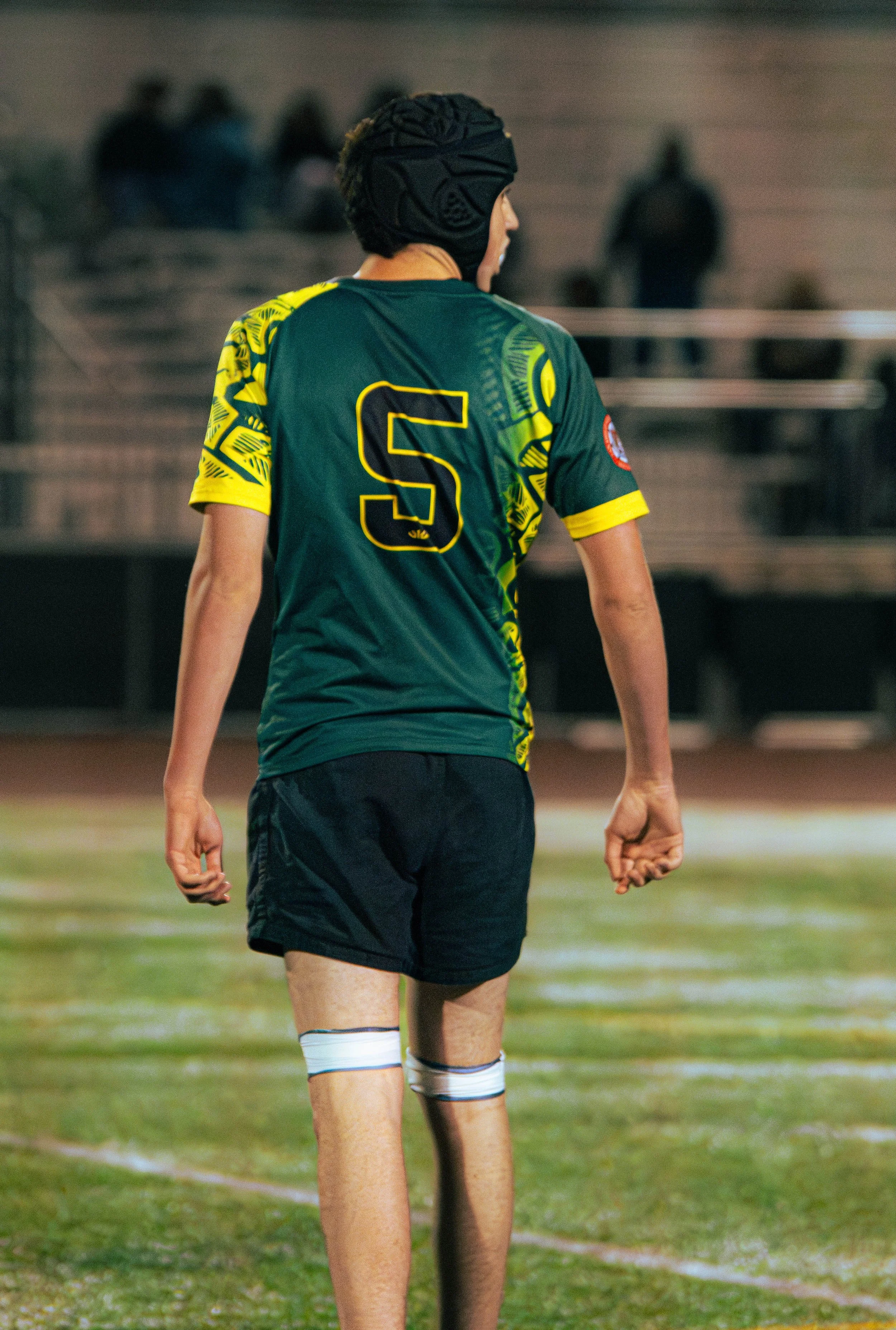 A rugby player wearing a green and yellow jersey with the letter 'S' on the back, black shorts, and a padded headgear, standing on a field at night.
