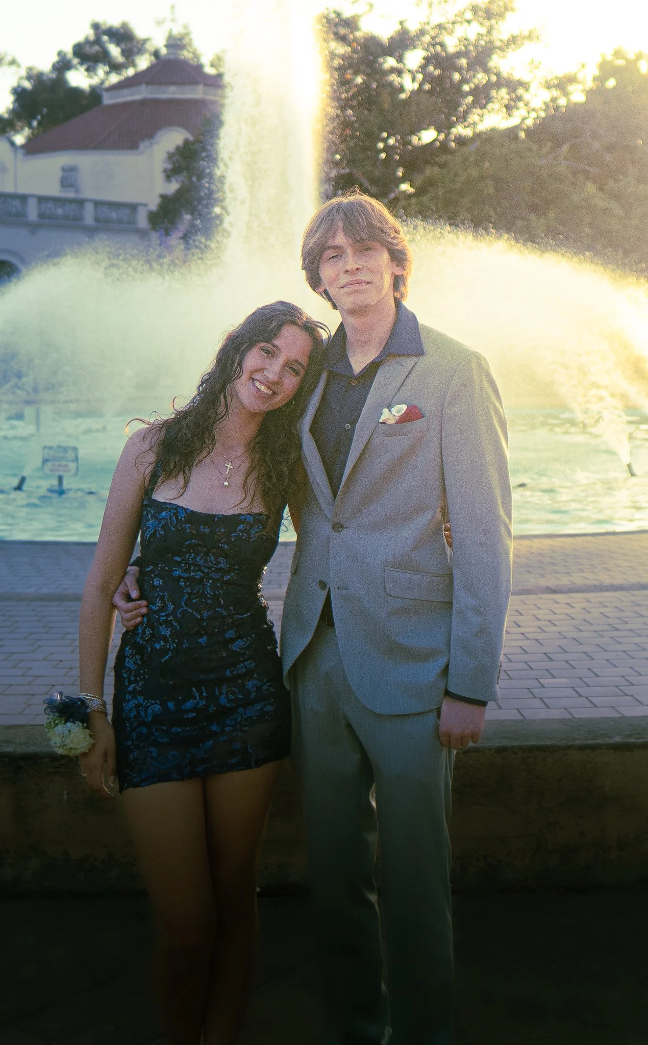 A woman and man stand close together in front of a fountain and trees on a sunny day. The woman is smiling, wearing a dark blue floral dress, and holding a small bouquet with a corsage on her wrist. The man is dressed in a light gray suit with a dark